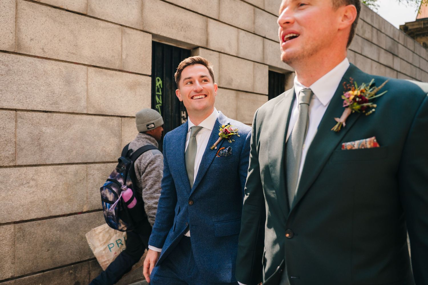 Two people in formal suits stand by a stone building wall sharing a candid laugh. Same sex wedding PHOTOGRAPHER Dublin