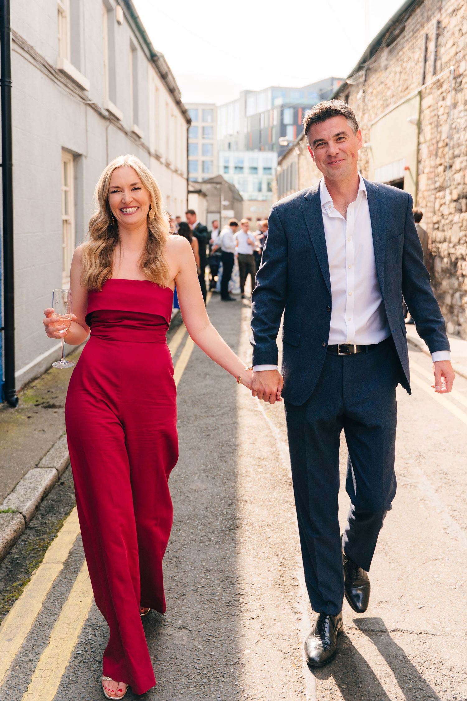 Wedding guests walking down a street in formal attire with a red dress standing out. Same sex wedding PHOTOGRAPHER Dublin