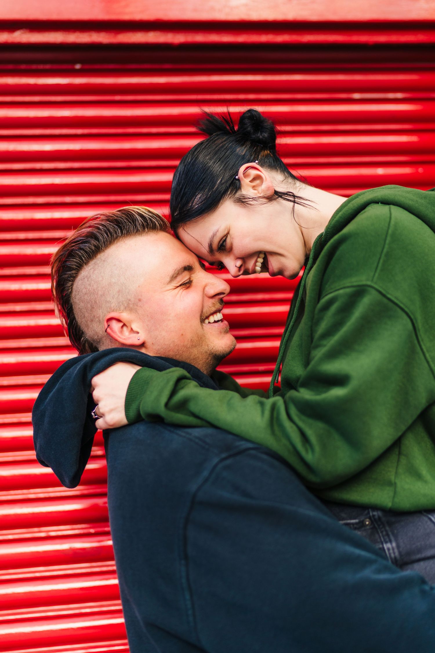 A couple laughing and embracing in front of a bright red corrugated metal wall. PHOTOGRAPHER Dublin Ireland