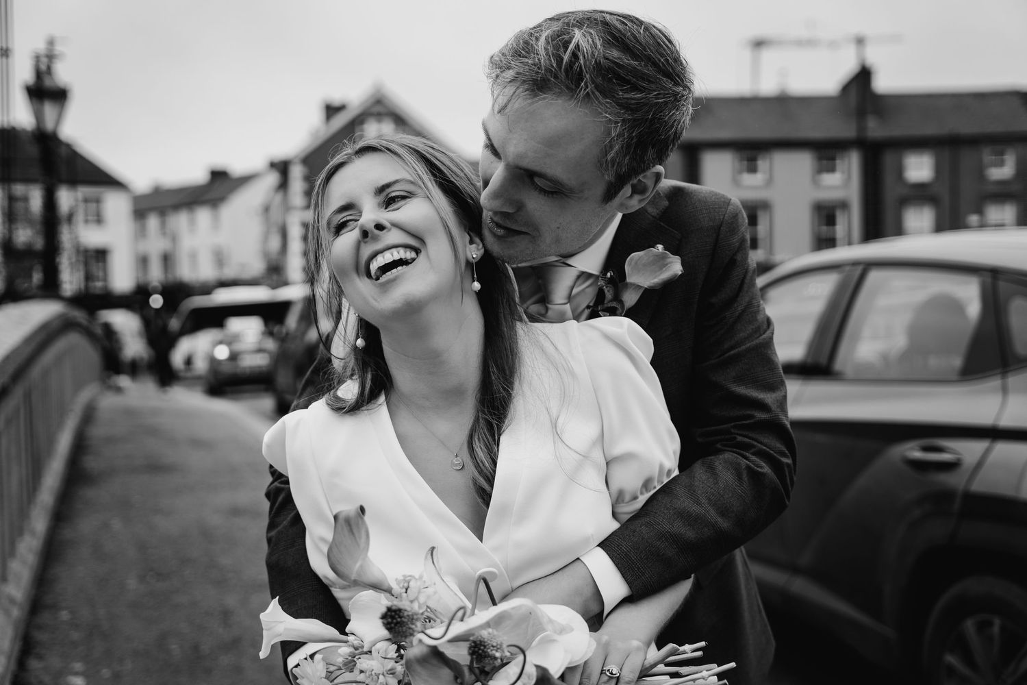 Black and white photo of a laughing bride and groom embracing on a street, capturing a joyful wedding moment.