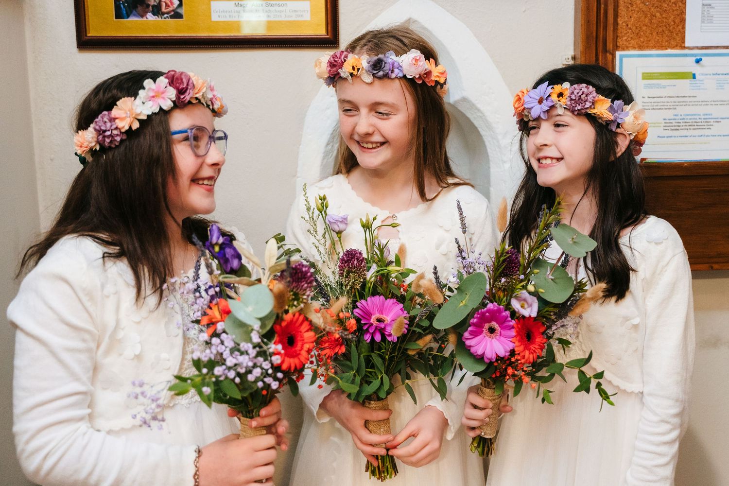 Three people in white dresses wearing floral crowns hold vibrant wedding bouquets while smiling together.