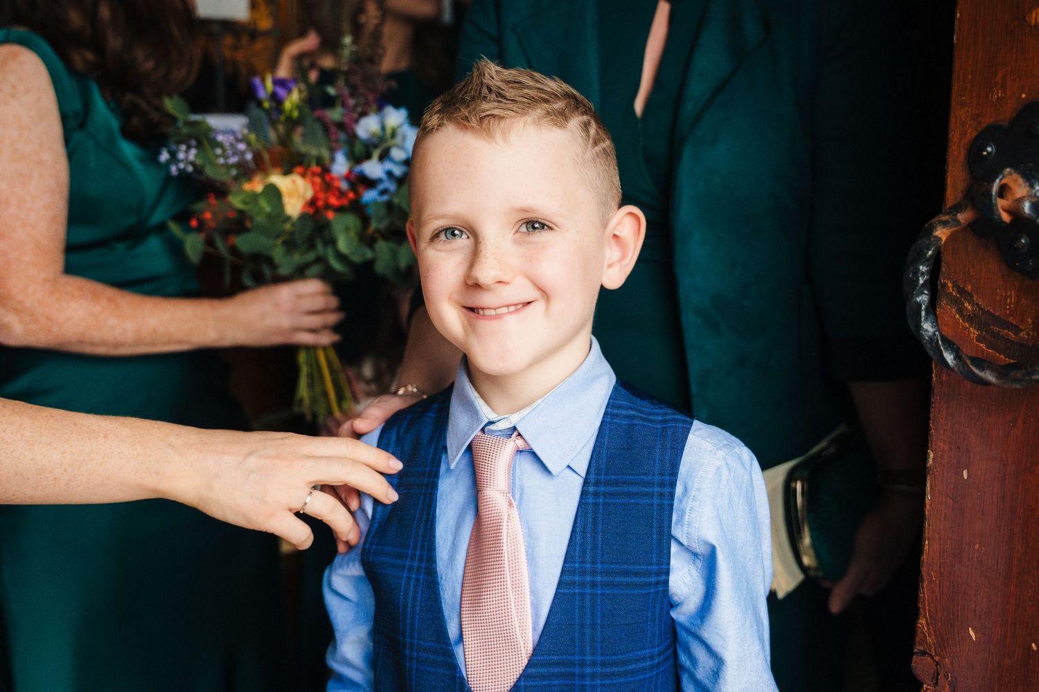 Young ring bearer in a blue vest and pink tie smiles at the camera.