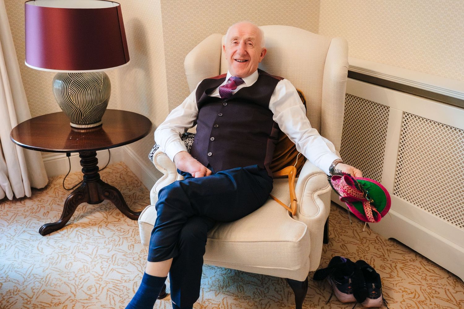 A cheerful elderly person in formal attire relaxes in a cream armchair next to a side table with lamp.