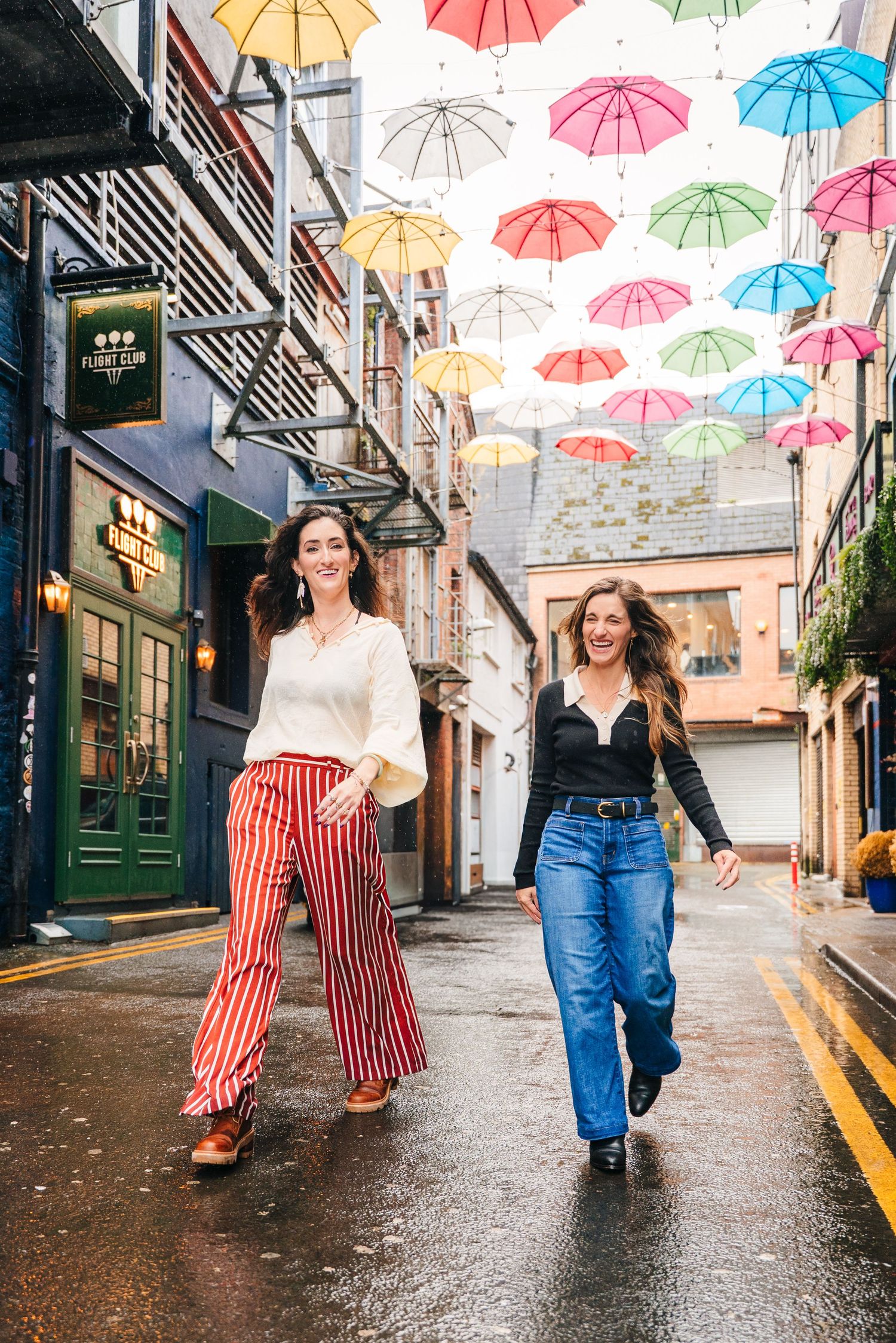 Two women walking under colorful umbrella art installation on rainy cobblestone street. professional headshots dublin photographer