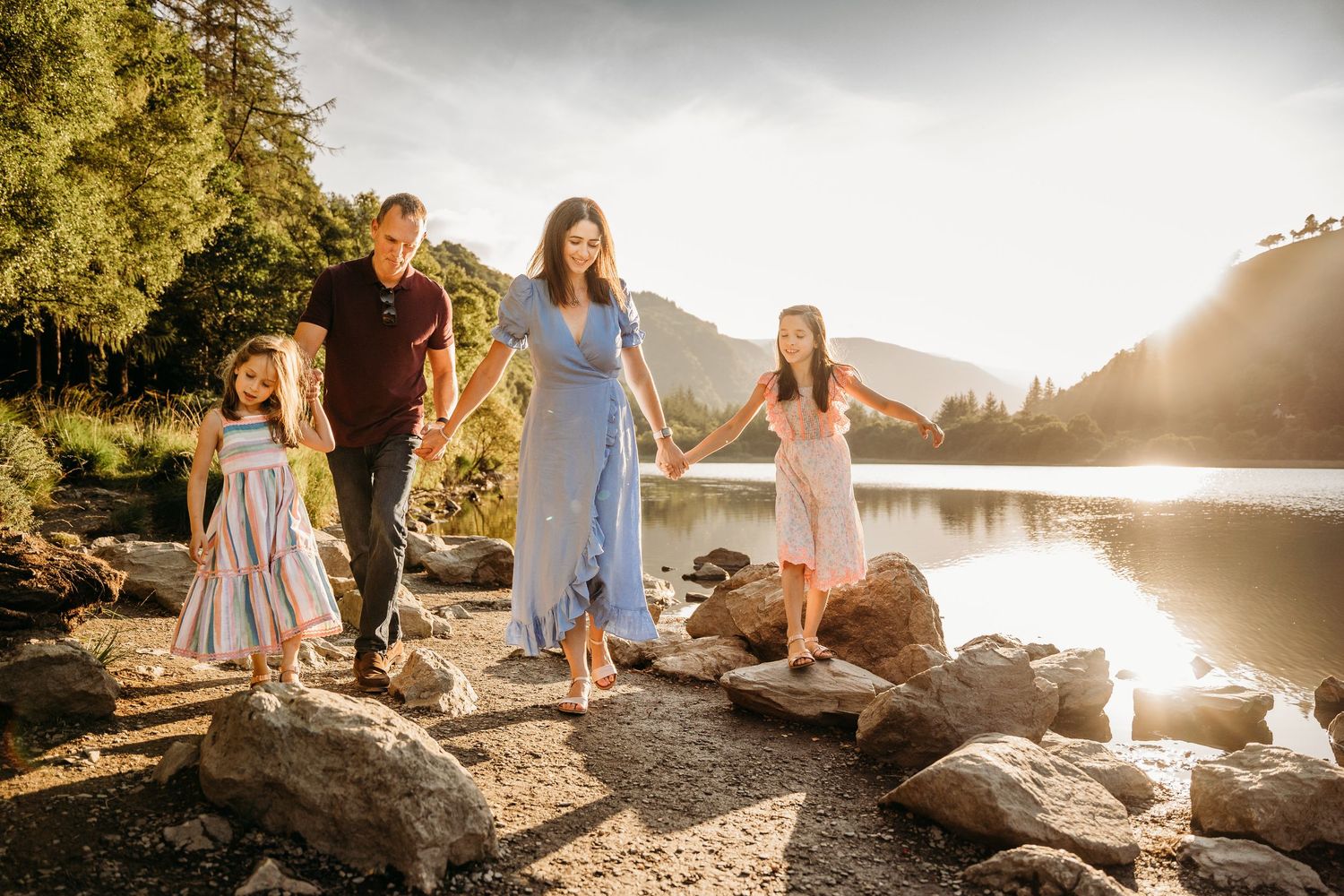 Family members sitting together on rocky shoreline with sun setting over mountain lake in background. family PHOTOGRAPHER Dublin