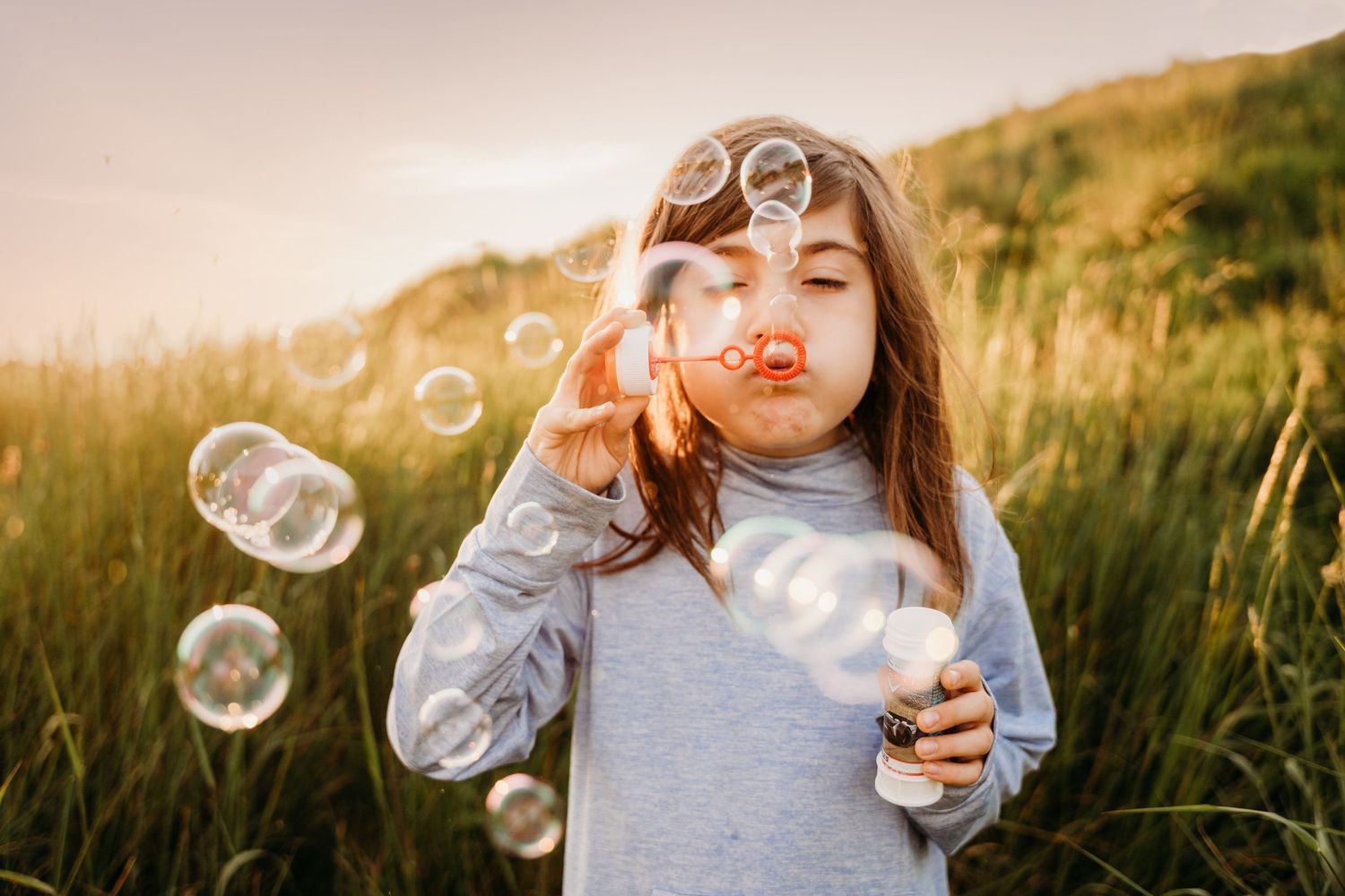 A child blowing soap bubbles in a sunlit meadow while wearing a gray sweater as golden light fills the scene. family PHOTOGRAPHER Dublin