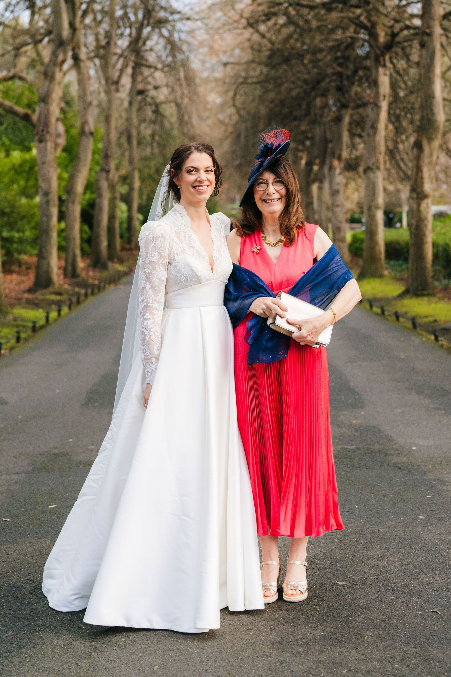 Two women walking down a tree-lined path, one in white wedding dress and one in red dress. Stephens green club dublin Wedding photographer