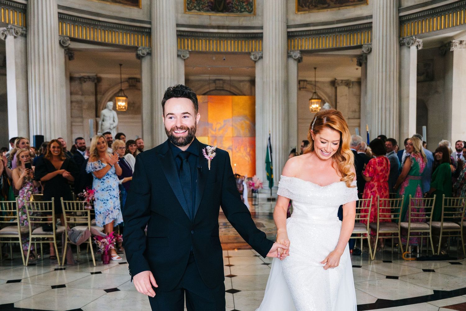 Wedding guests celebrate in an elegant columned venue with gold chairs and decorative details.  city hall dublin wedding photos