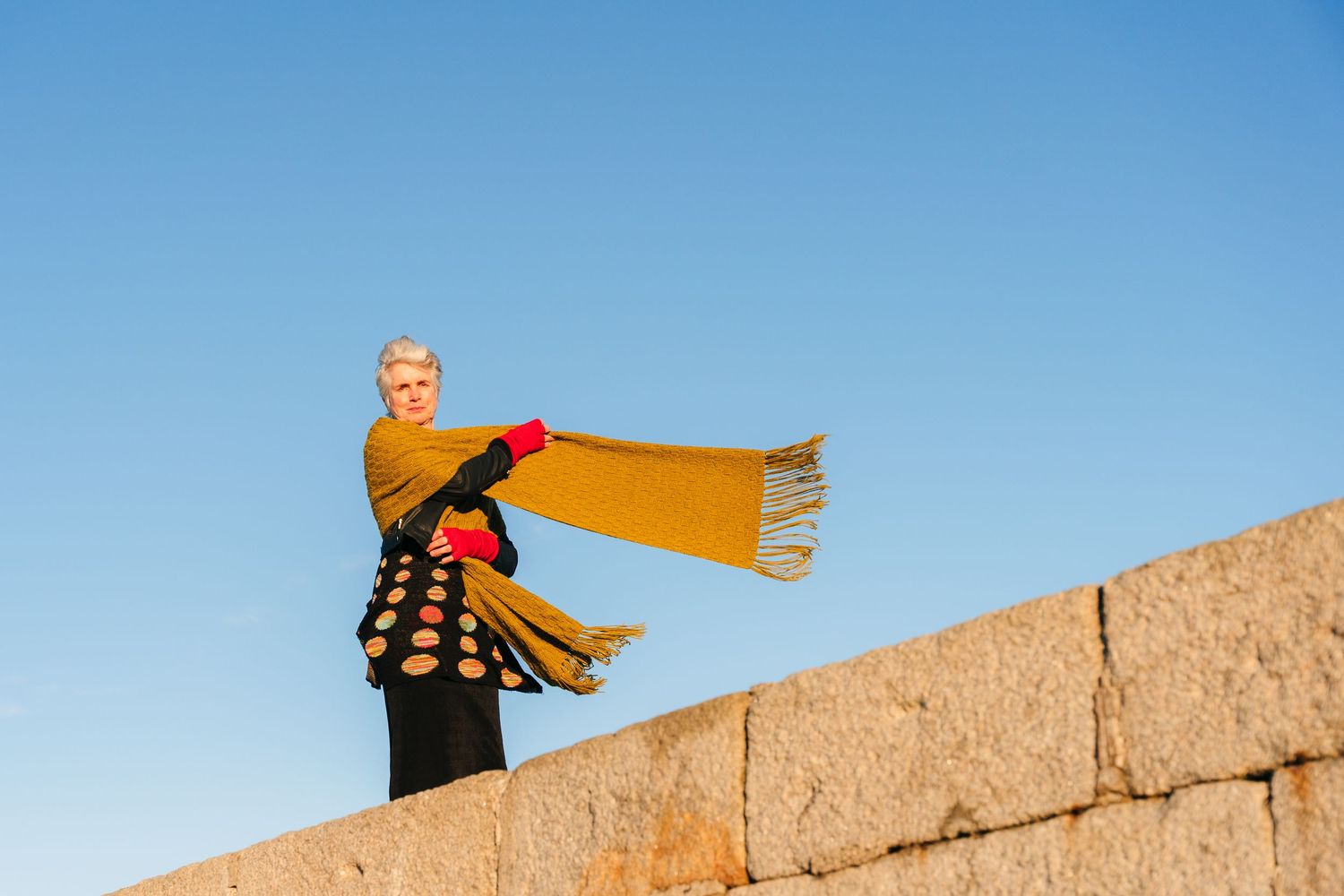A person wearing a yellow sweater stands on a stone wall with a scarf blowing in the wind. Dublin photographer professional headshots