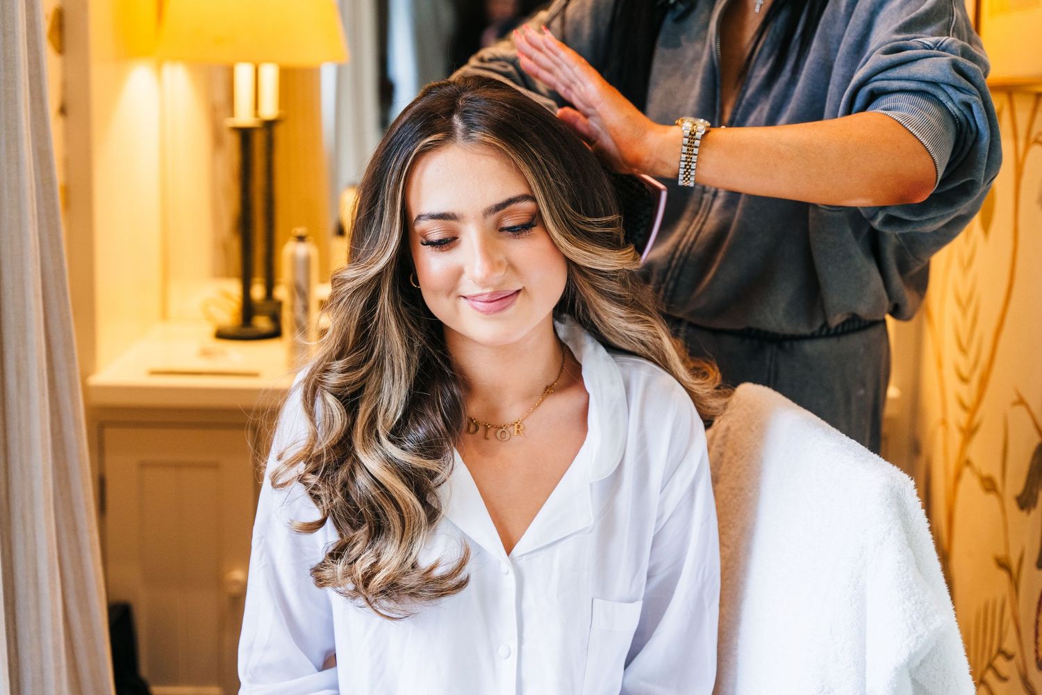 A hairstylist styles wavy brown hair in room with warm lighting and white robe. Virginia Park Lodge Wedding Photographer Dublin Meath Irelan