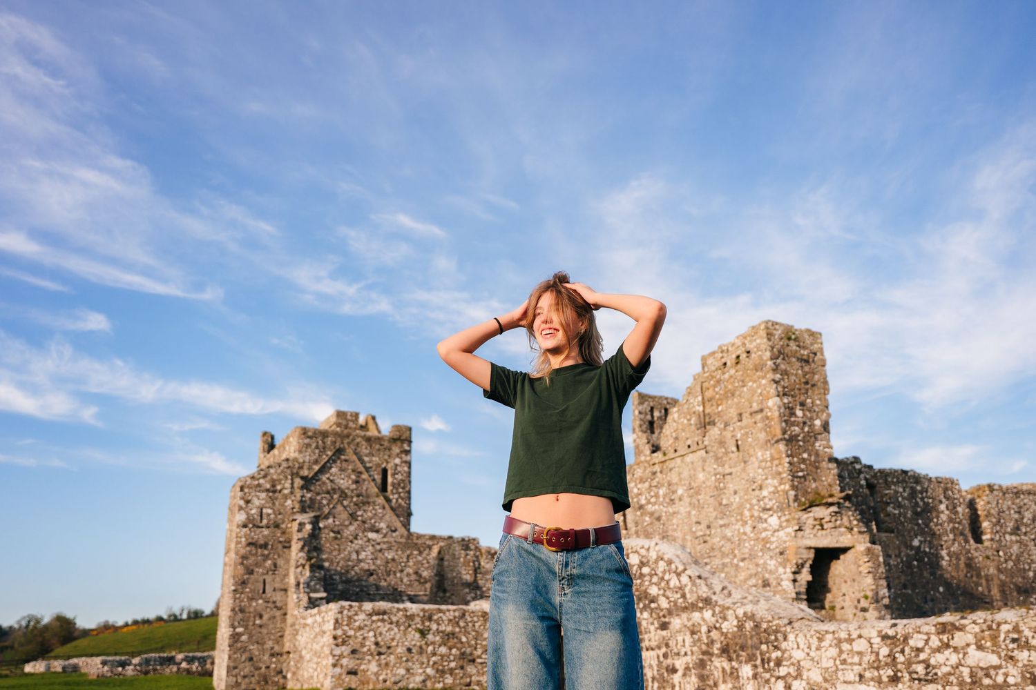 Person in jeans and cropped top stands on ancient stone ruins against blue sky with wispy clouds. Photographer Ireland dublin