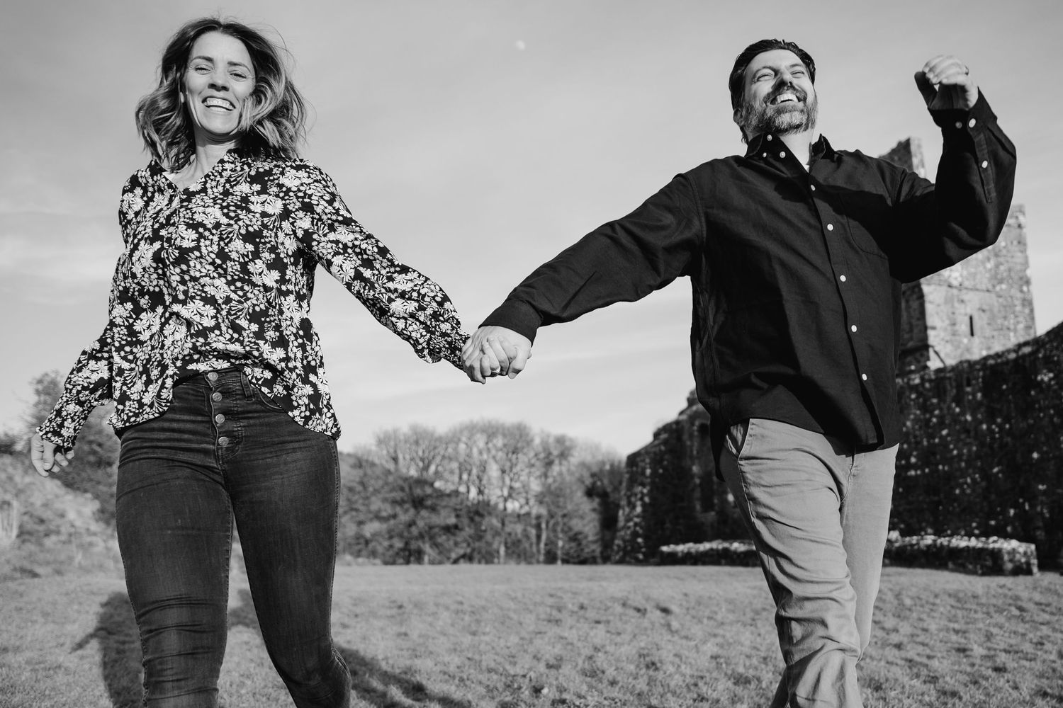 Two people hold hands while joyfully jumping together in a field, captured in black and white. Photographer Ireland dublin
