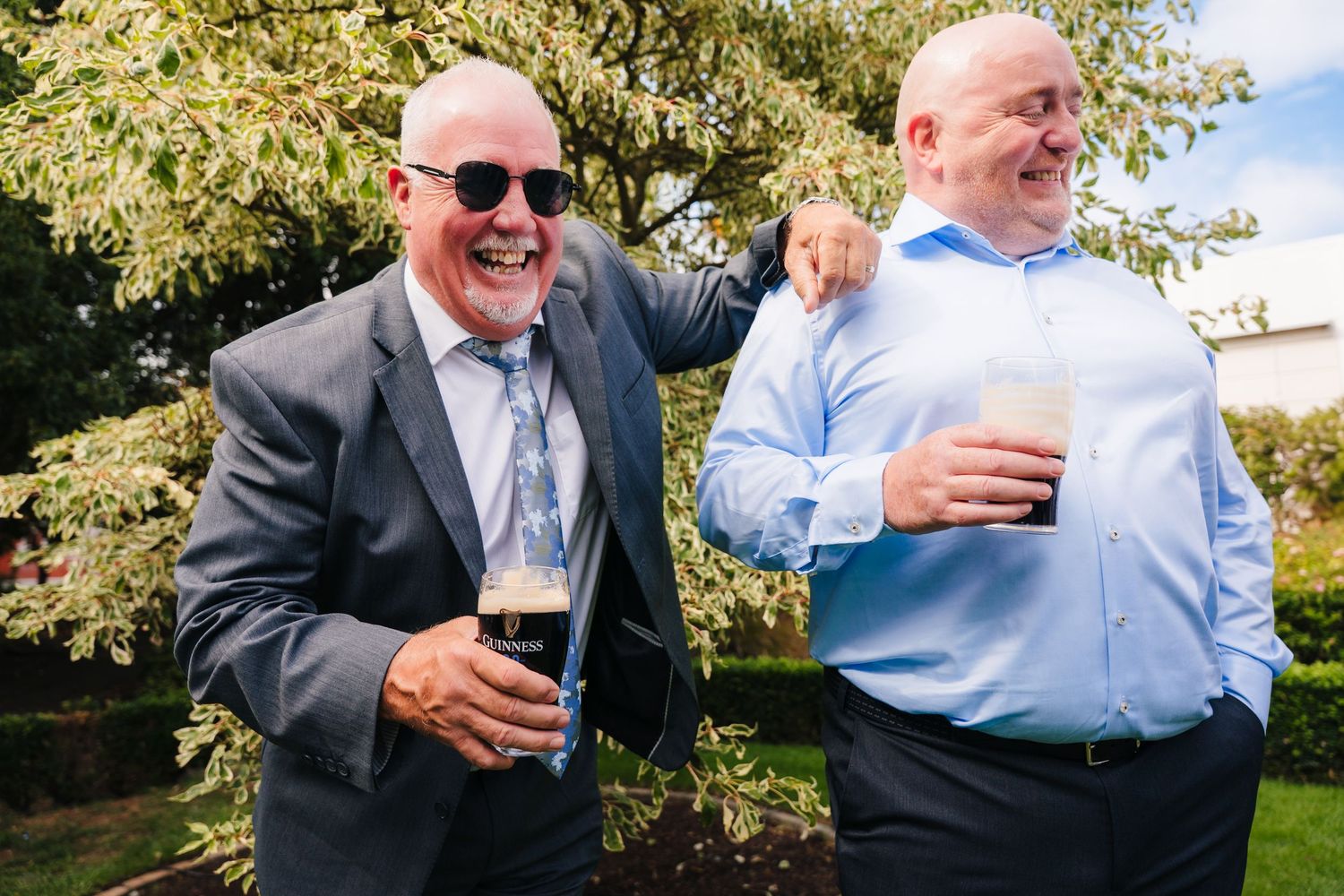 Guests sharing drinks and laughs outdoors at a formal event under green foliage. Portmarnock Hotel Wedding PHOTOGRAPHER Dublin