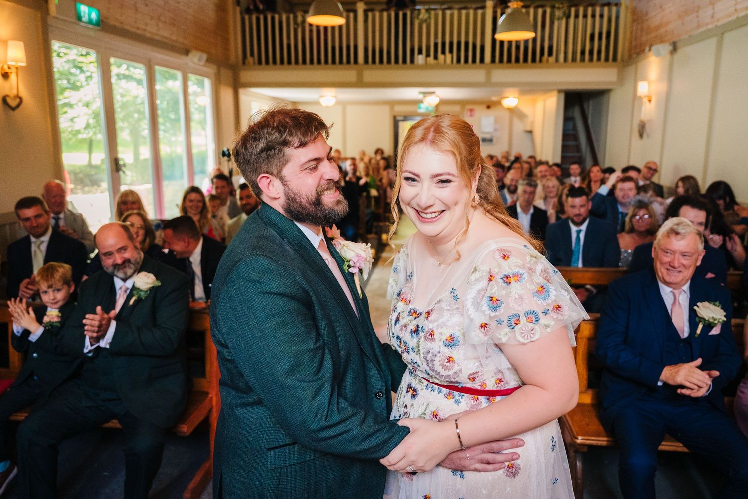 A couple shares a joyful moment during their wedding ceremony while guests look on in a chapel setting. . Village Barn Tyrrellspass wedding