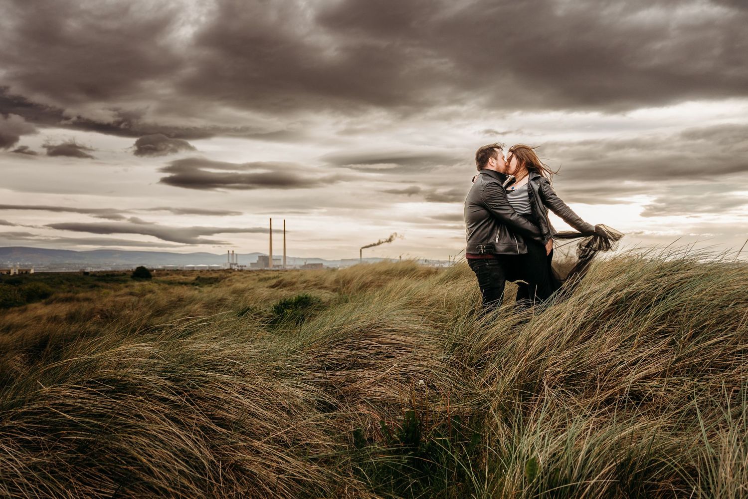 Panoramic coastal landscape with two figures embracing among windswept beach grass under moody skies. engagement PHOTOGRAPHER Dublin