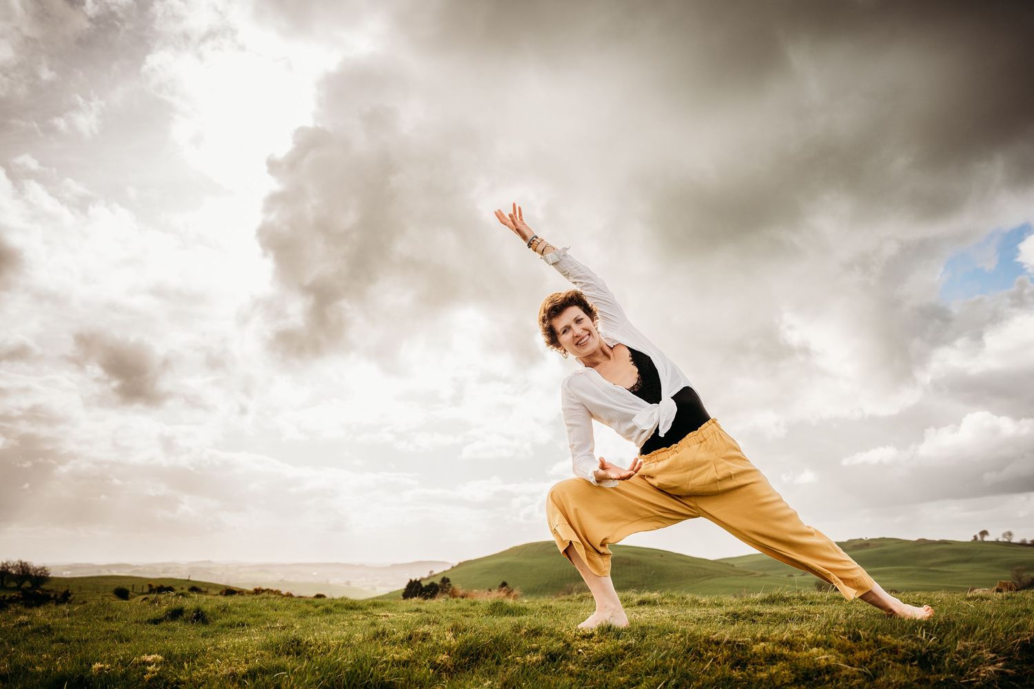 Dancer in white top and yellow pants strikes dynamic pose against dramatic cloudy sky over green hillside. Portrait PHOTOGRAPHER Dublin