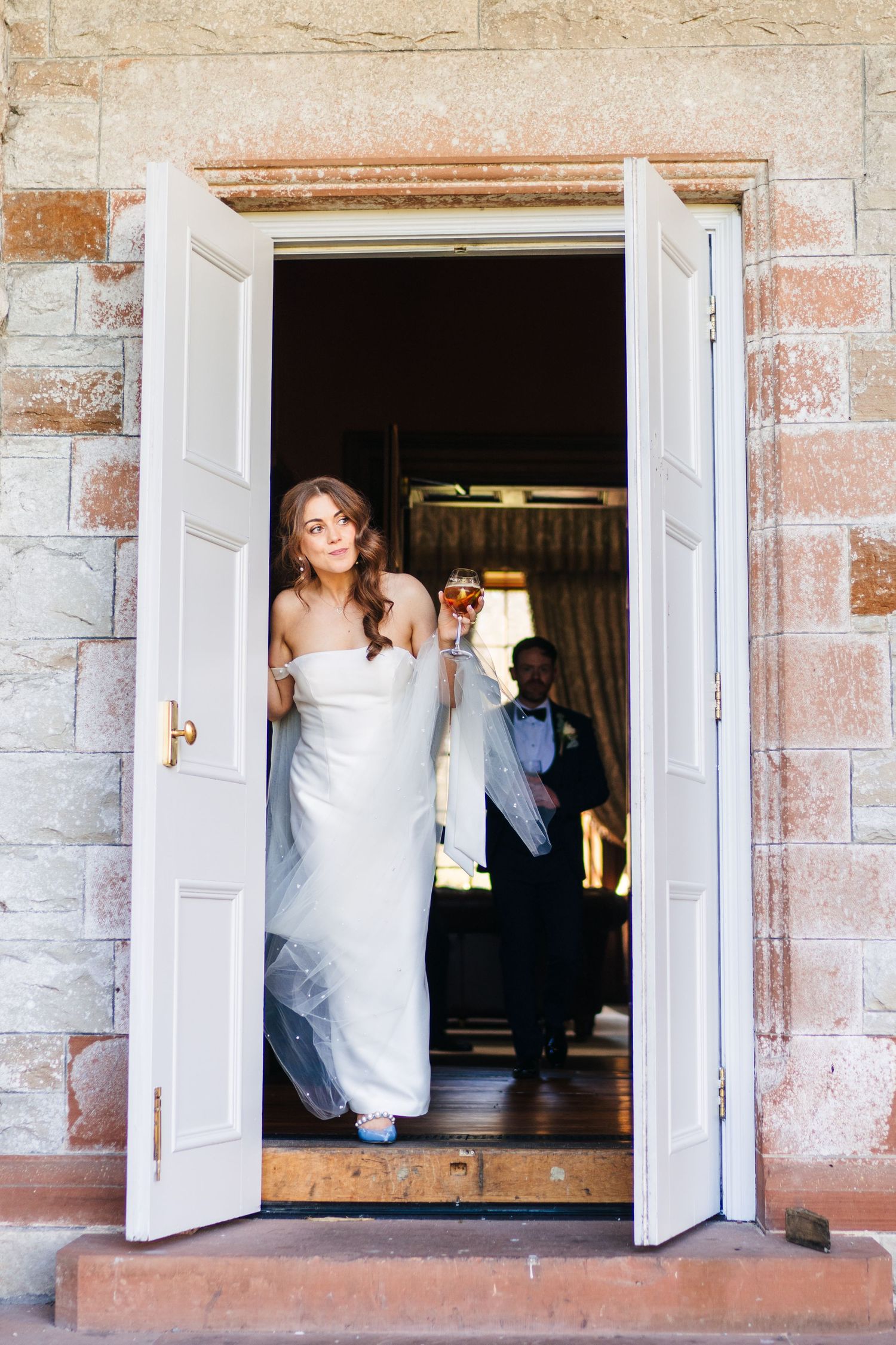 Castle Leslie Wedding PhotSomeone in an elegant white wedding dress stands in a doorway of a rustic stone building with white double doors.