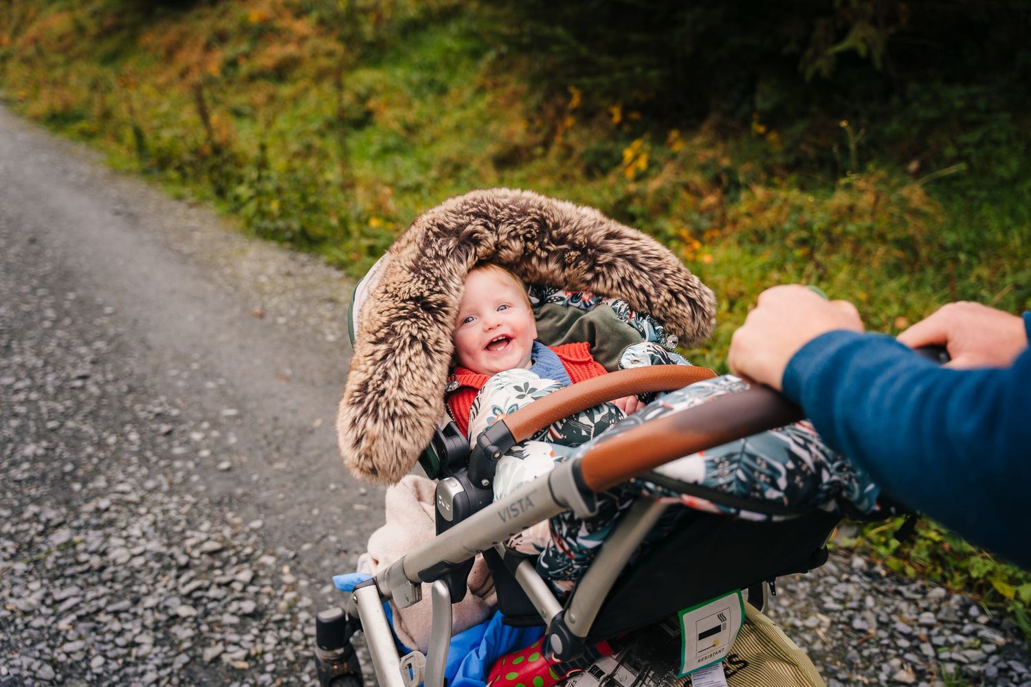 Family Photographer Dublin Ireland Baby bundled in a cozy fur-trimmed hood sits in a stroller on a rural path.