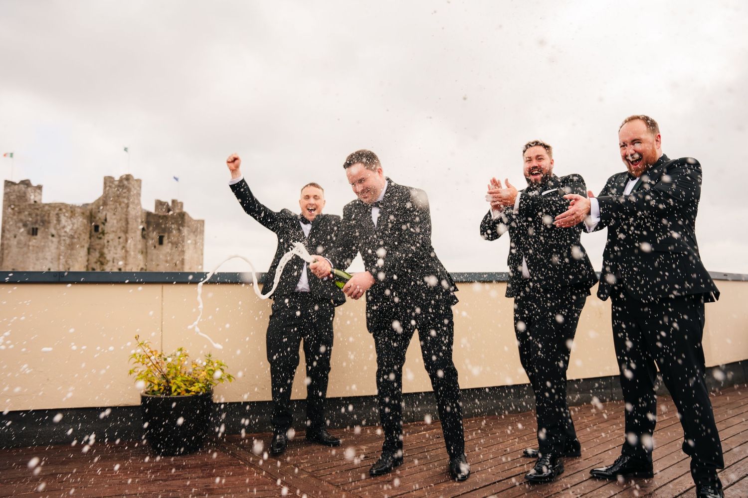 Wedding party in black suits celebrating with confetti on a rooftop with a castle in the background. Wedding Photography Trim Castle Ireland