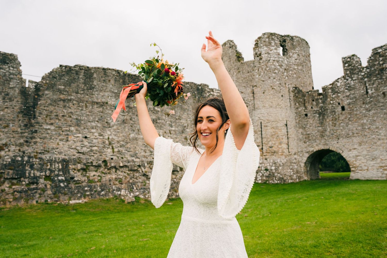 A person in a wedding dress celebrates with raised arms holding a bouquet in front of castle ruins.Wedding Photography Trim Castle Ireland