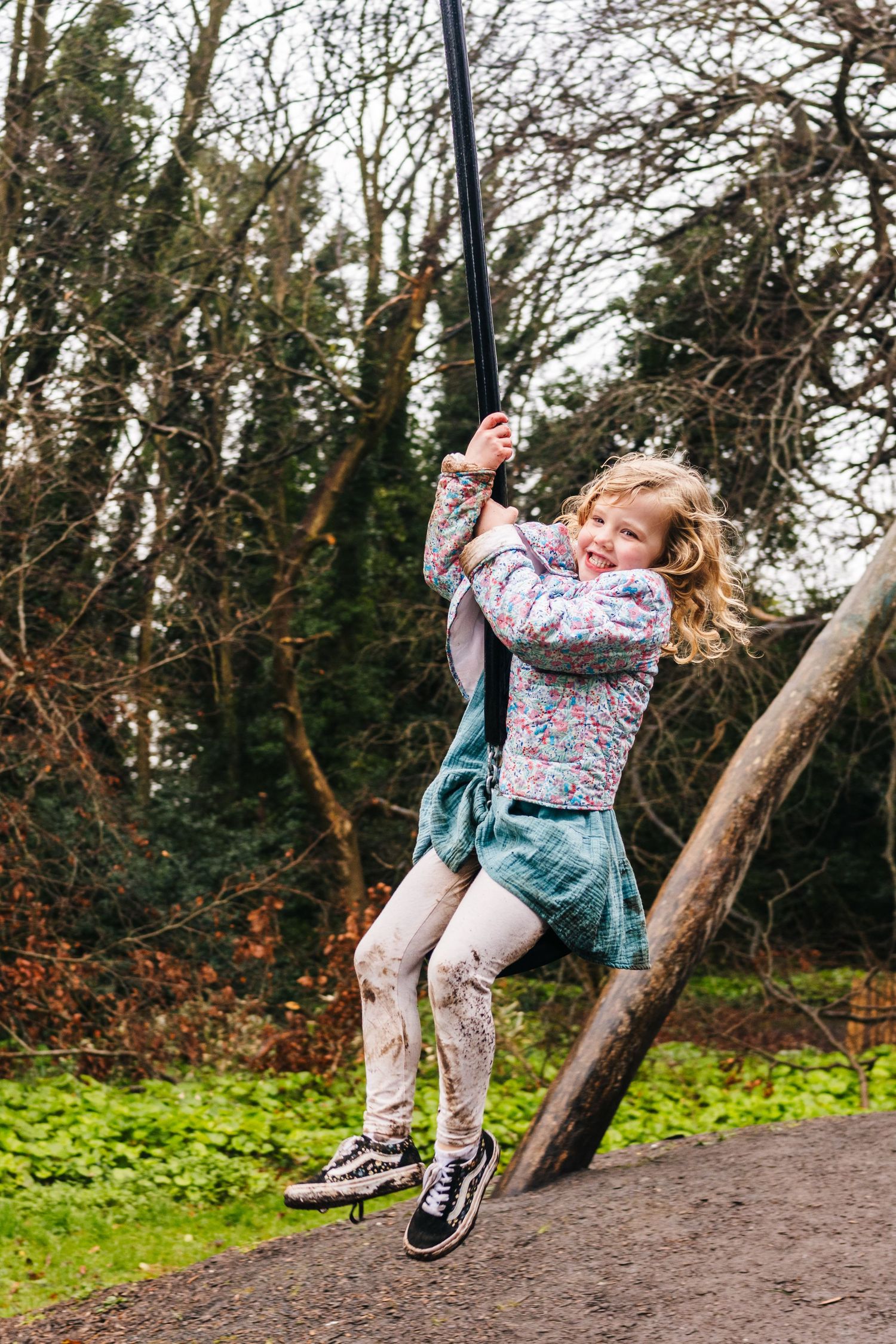 Family photographer Dublin Ireland  A child in a floral top and denim skirt swings joyfully on a rope swing in a wooded outdoor setting.