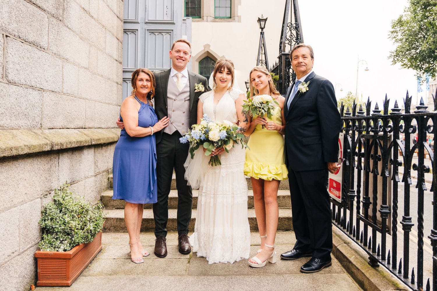 Wedding party poses for formal photo outside stone building with decorative iron fence. PHOTOGRAPHER Dublin Ireland Wedding