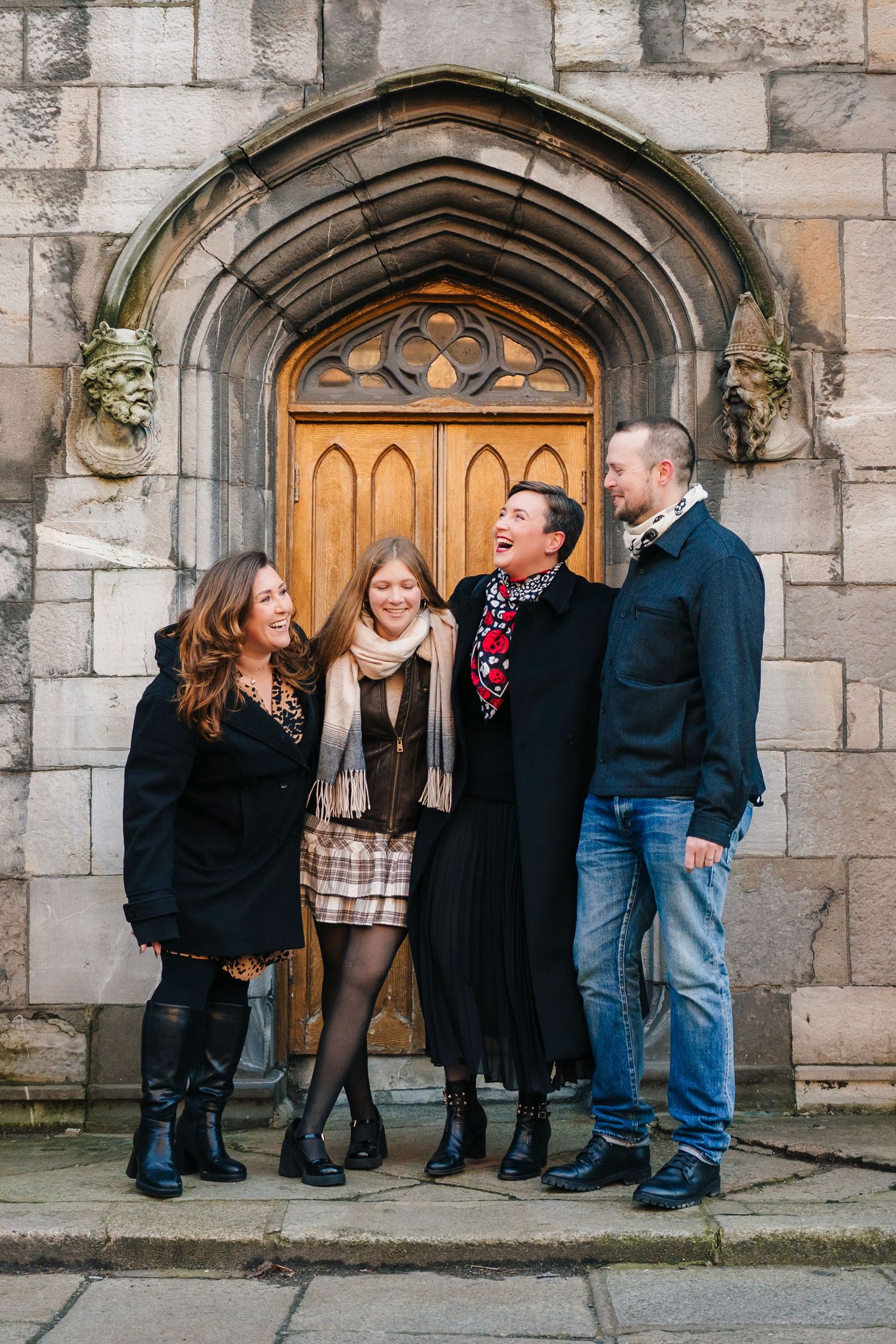 A group of people stand together and laugh in front of an ornate stone church entrance with wooden arched doors. family photographer dublin
