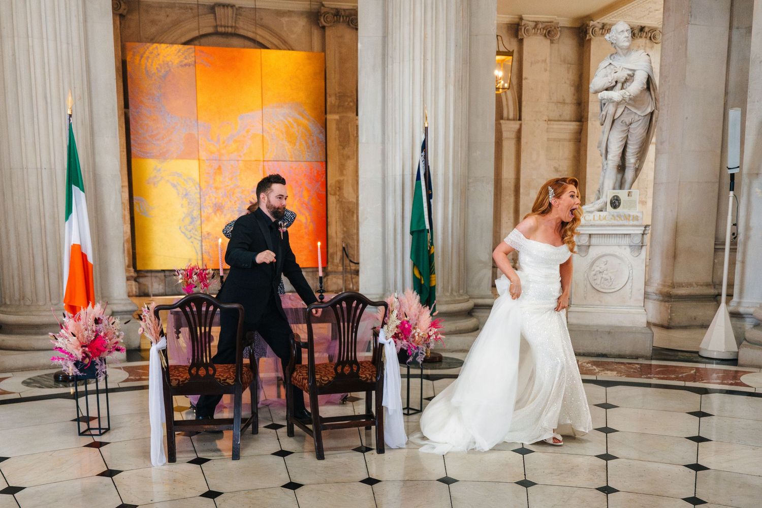Wedding celebration in grand hall with white columns and orange-lit walls featuring Irish flags.  city hall dublin wedding photos