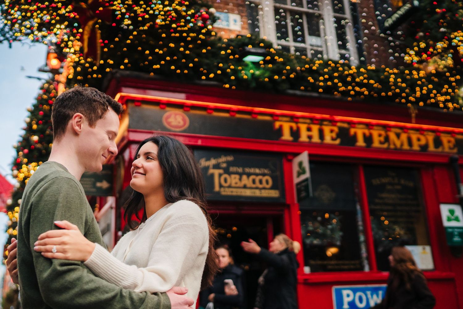 A couple embraces outside The Temple Bar in Dublin with festive lights twinkling overhead. Surprise proposal dublin photography