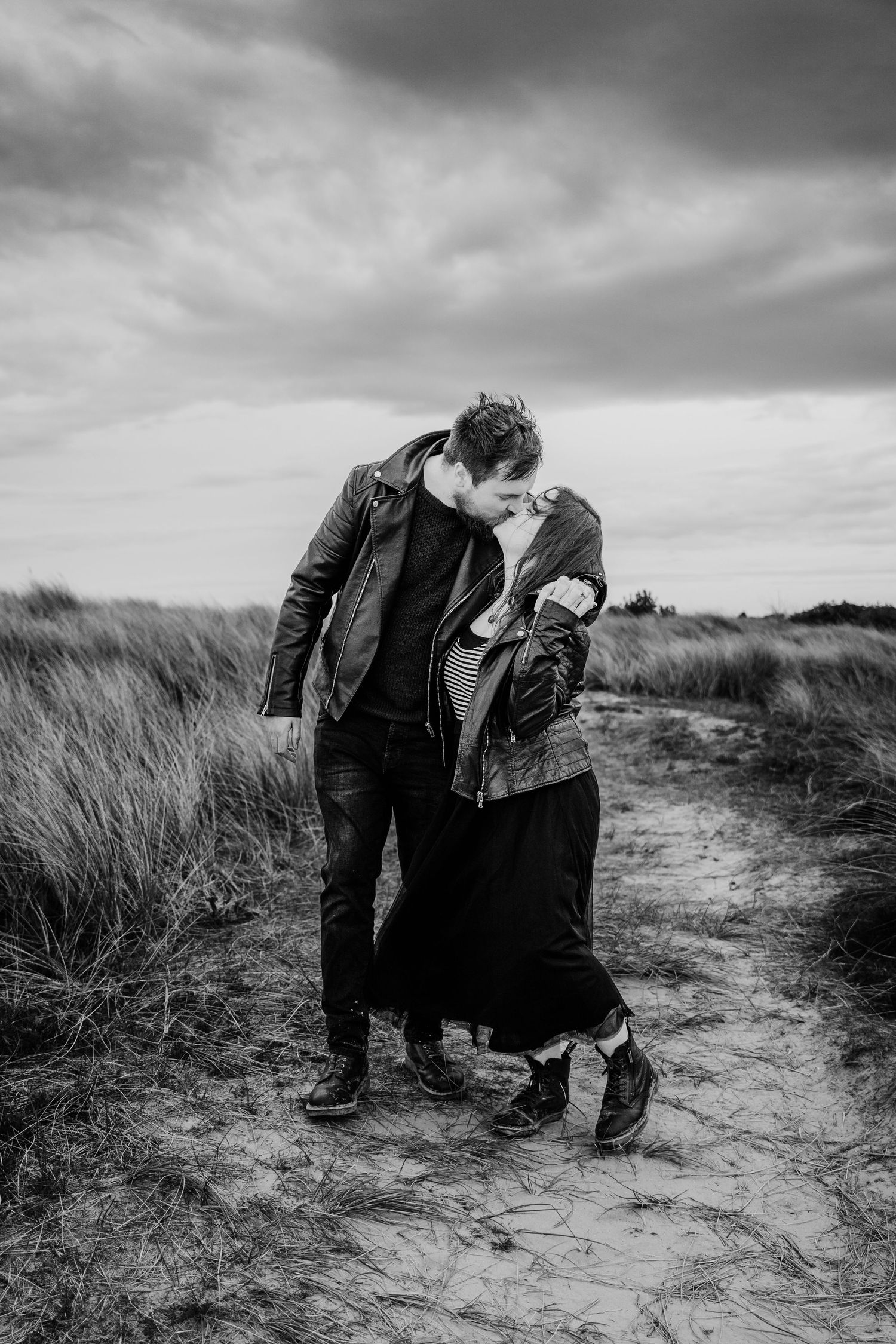 Dramatic black and white photo of a couple in leather jackets on a windswept beach path with stormy skies. engagement PHOTOGRAPHER Dublin