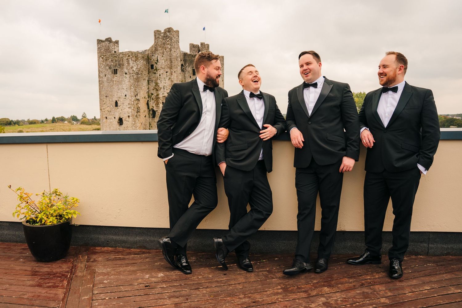 A group of groomsmen in black tuxedos share a laugh while standing against a backdrop of a historic castle. Wedding Photography Trim Castle