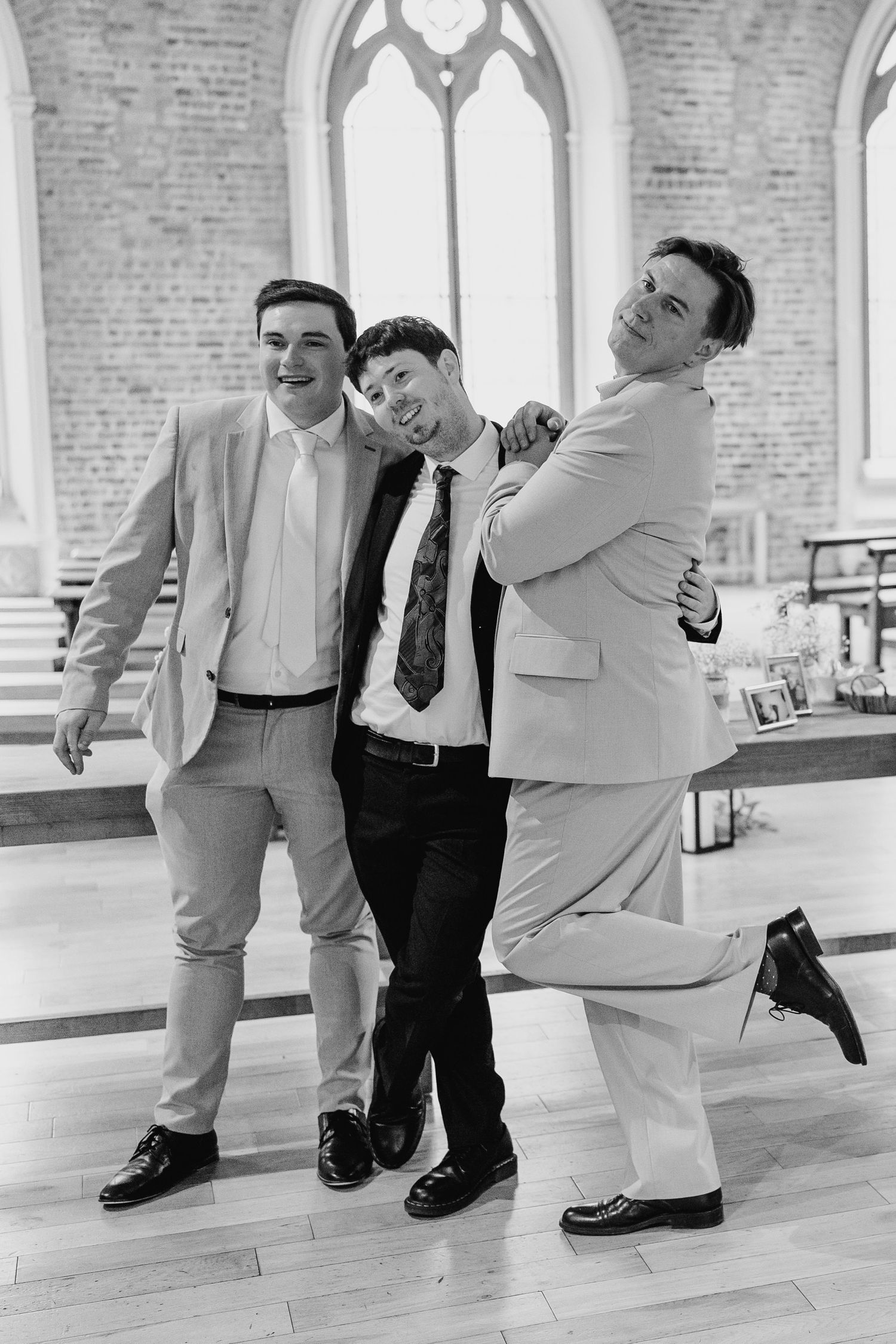Three groomsmen strike a playful pose in a church venue with gothic windows and exposed brick walls. PHOTOGRAPHER Dublin Ireland Wedding