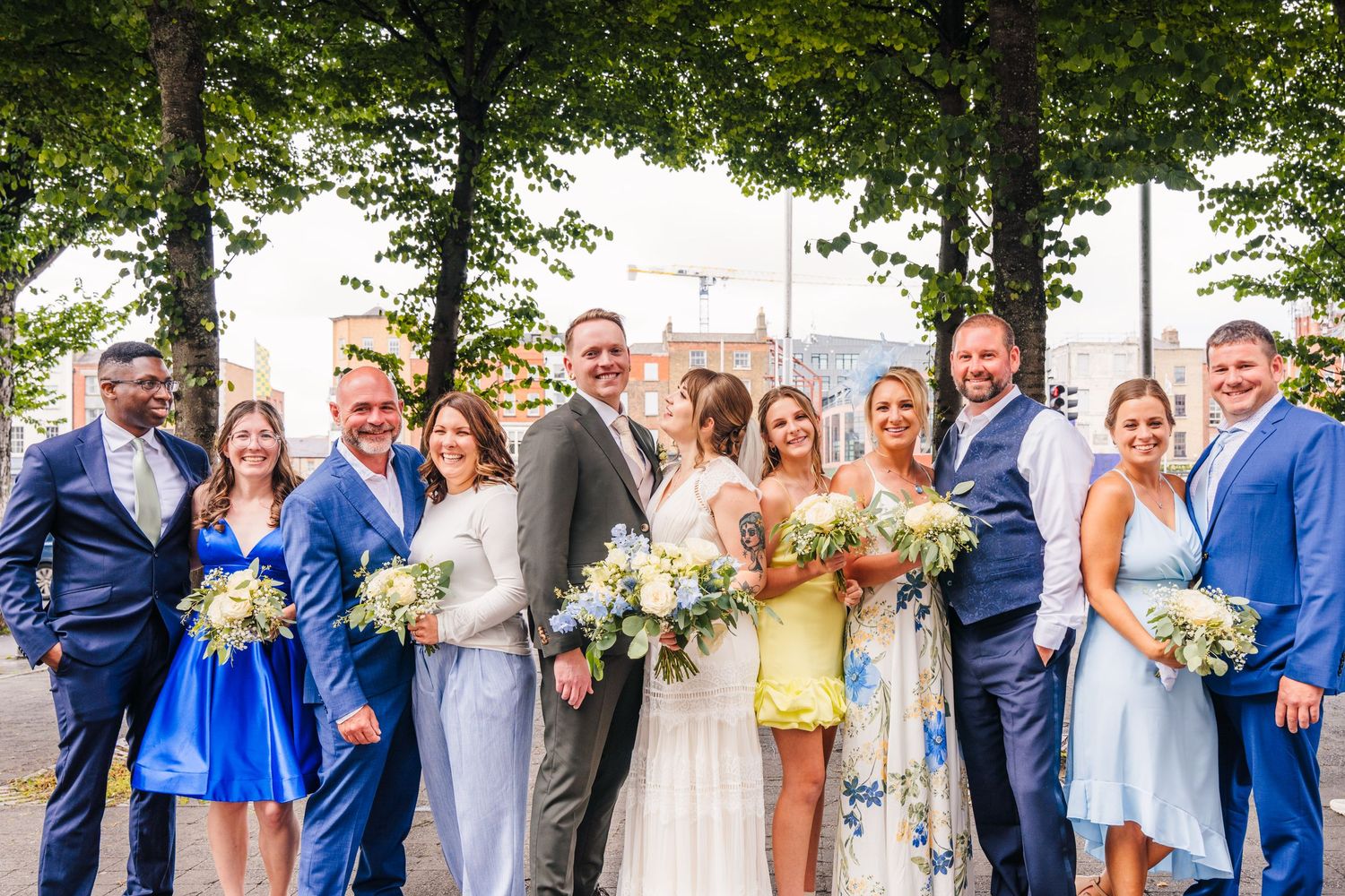 Wedding party in blue and neutral attire poses together for a formal group photo under trees. PHOTOGRAPHER Dublin Ireland Wedding