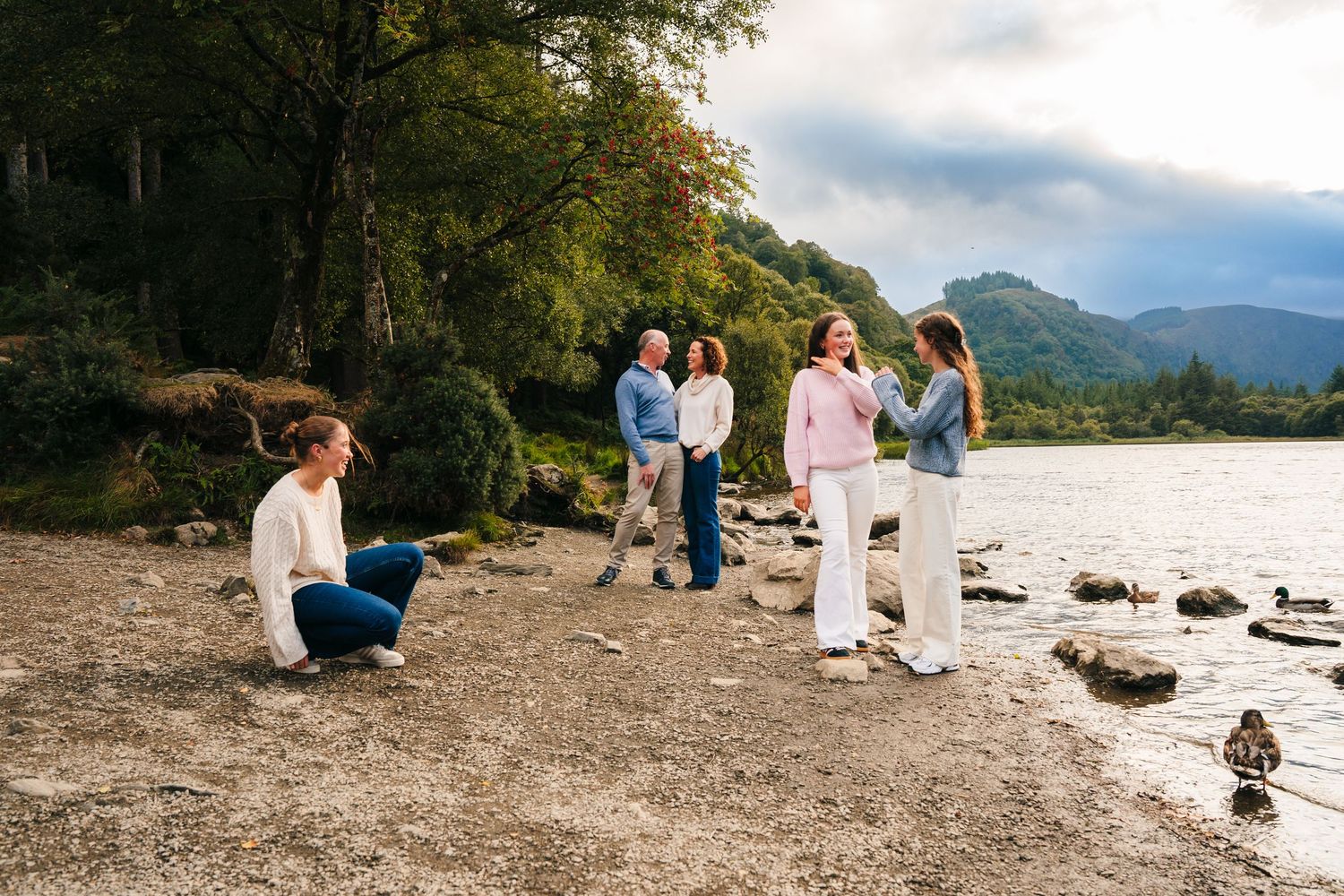 Family photographer Ireland Dublin Couple enjoys a peaceful moment on a rocky lakeshore surrounded by mountains during sunset.