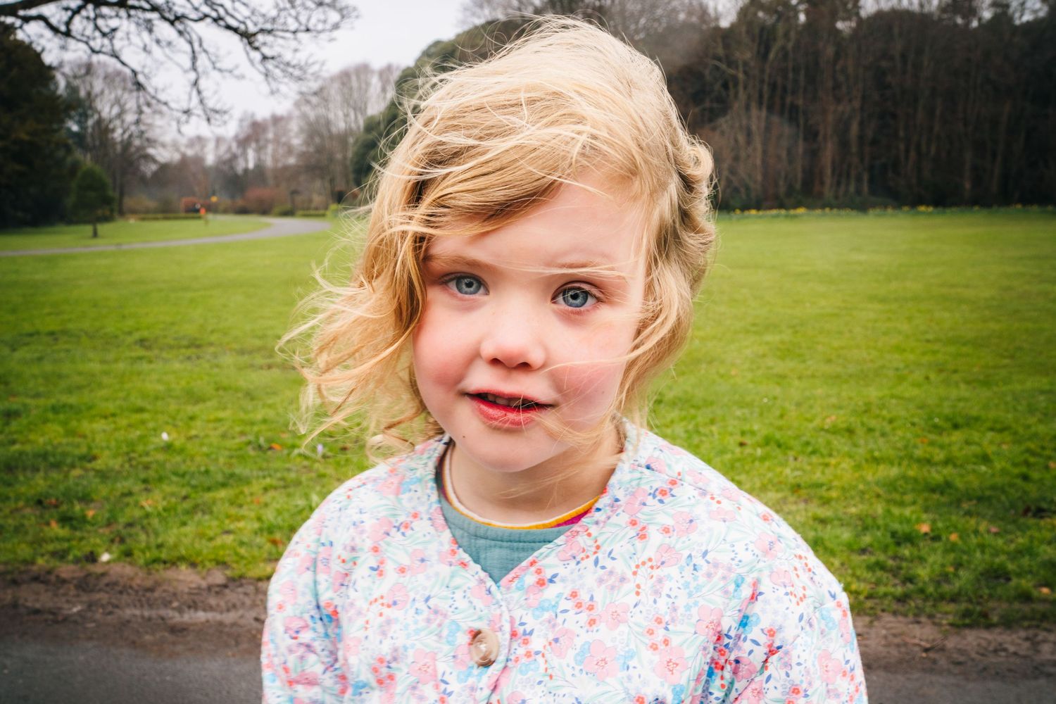 Young child in floral print clothing stands in a park with green grass and trees in the background. Family photographer Dublin Ireland
