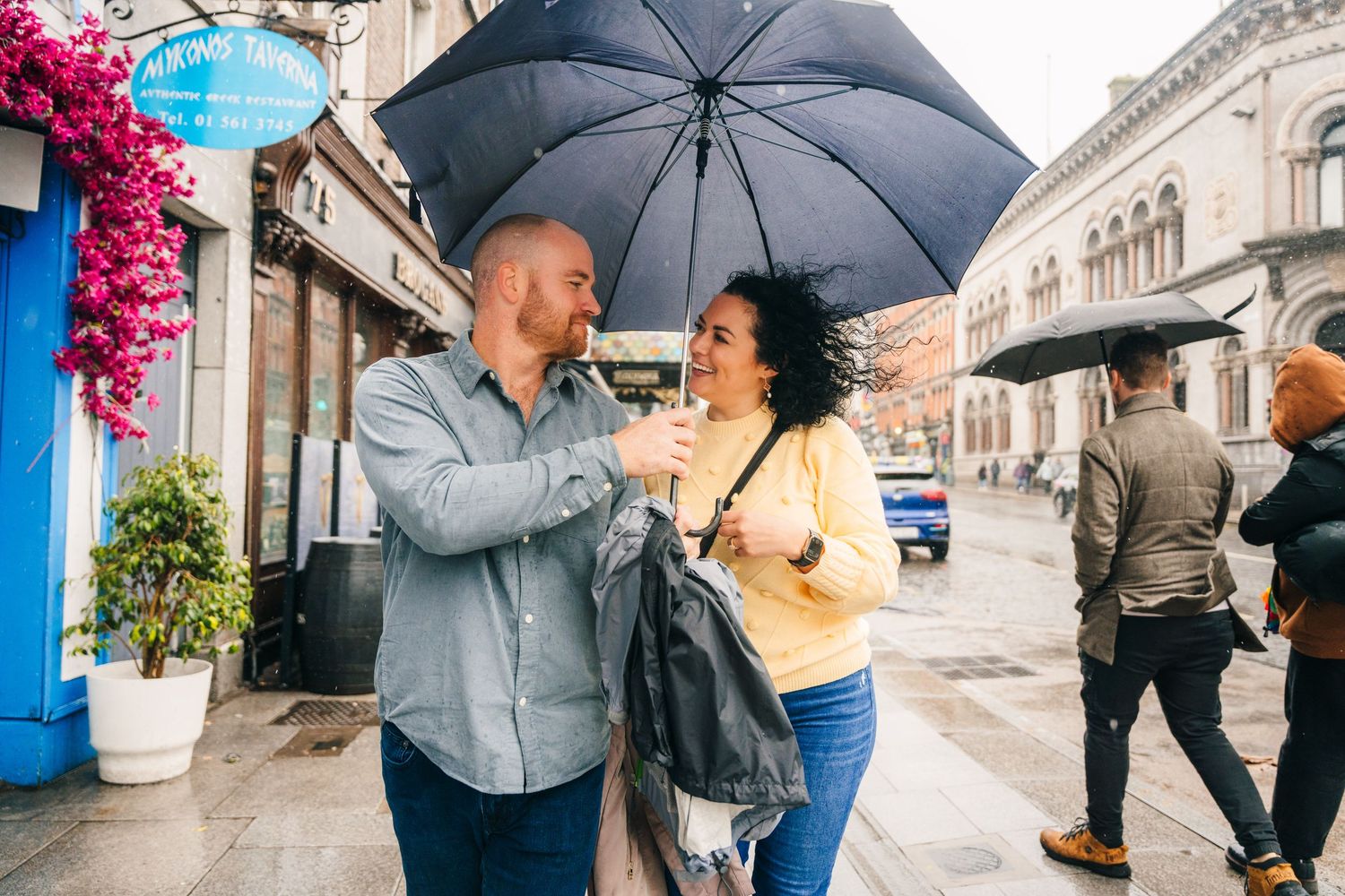 Two people share a black umbrella while walking and laughing together on a rainy city street with storefronts. Proposal Dublin Photographer