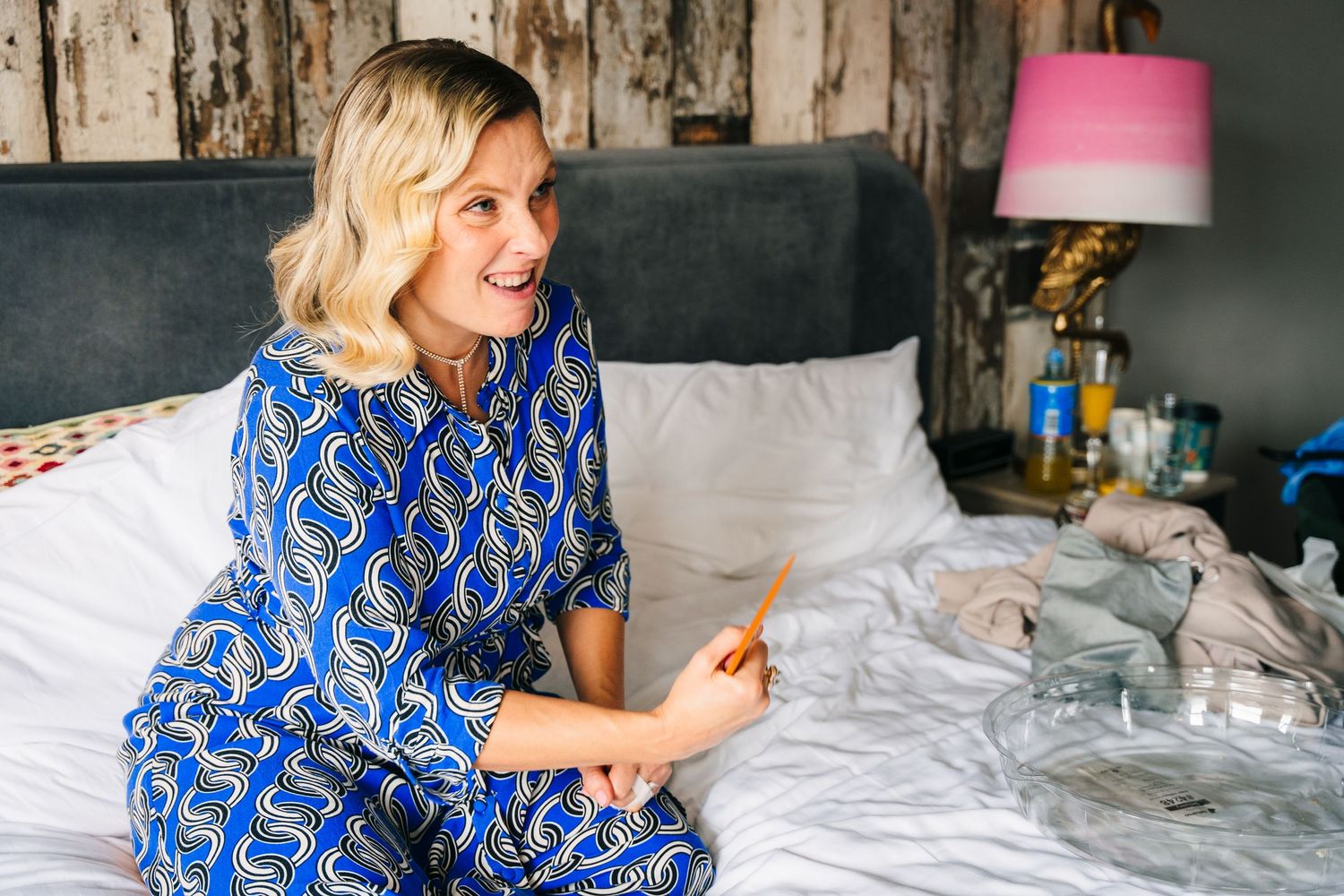 Someone in a blue patterned dress sits on a white bed with pillows and a pink lamp in the background. House Dublin Wedding Photographer