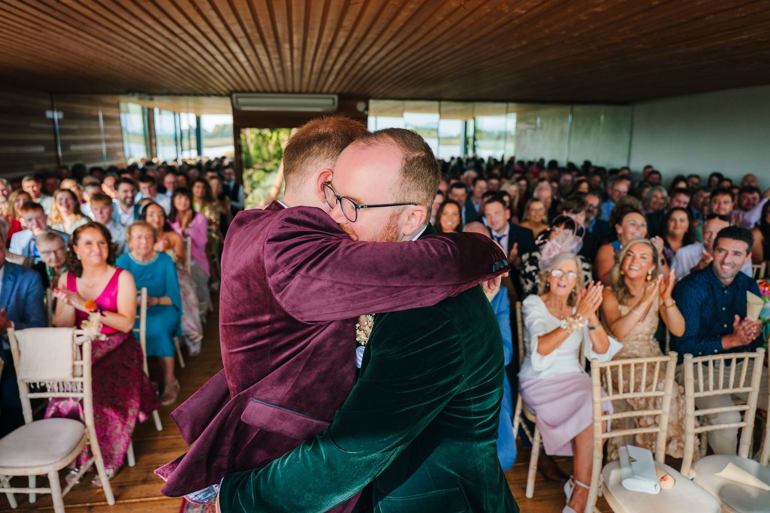 Wedding guests applaud emotionally during a heartfelt embrace at an indoor reception venue with wooden ceiling. LGBTQ Ashley Park Wedding Ph