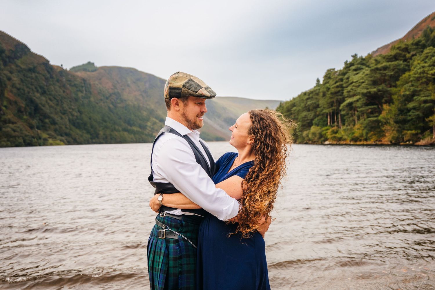 Couple embracing by a scenic lake surrounded by mountains in Scotland.Family photogrAPHER DUBLIN