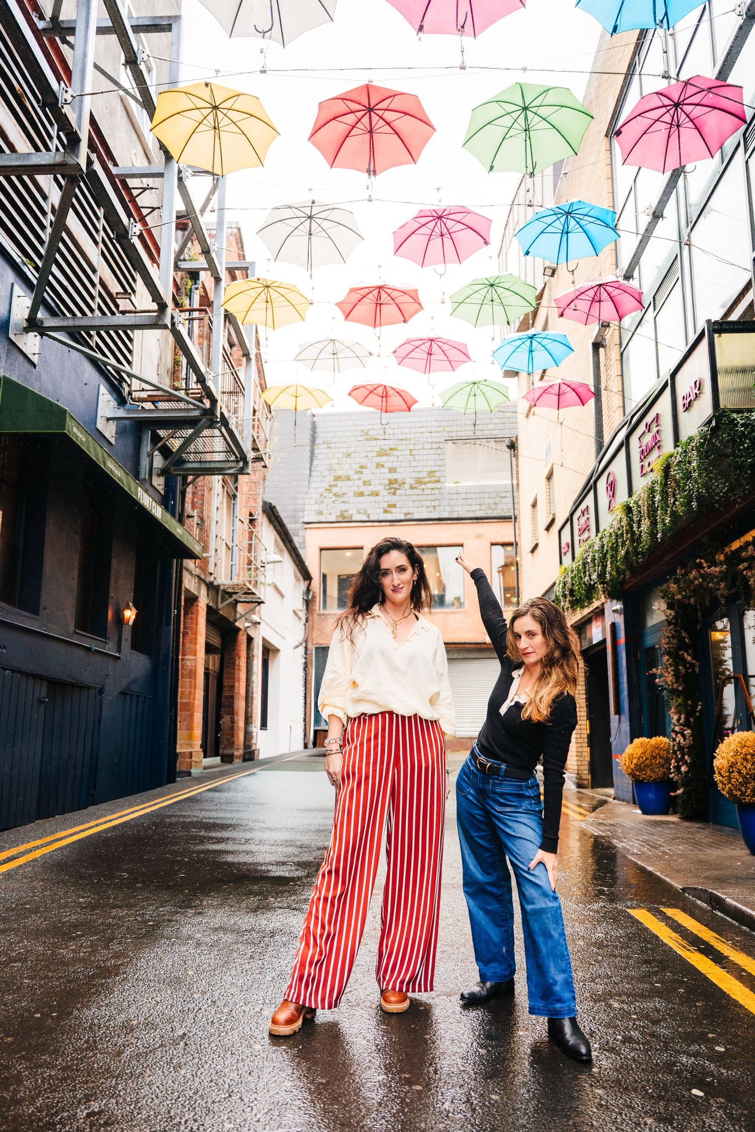 Two friends dancing under colorful suspended umbrellas in an urban alleyway on a rainy day. professional headshots dublin photographer