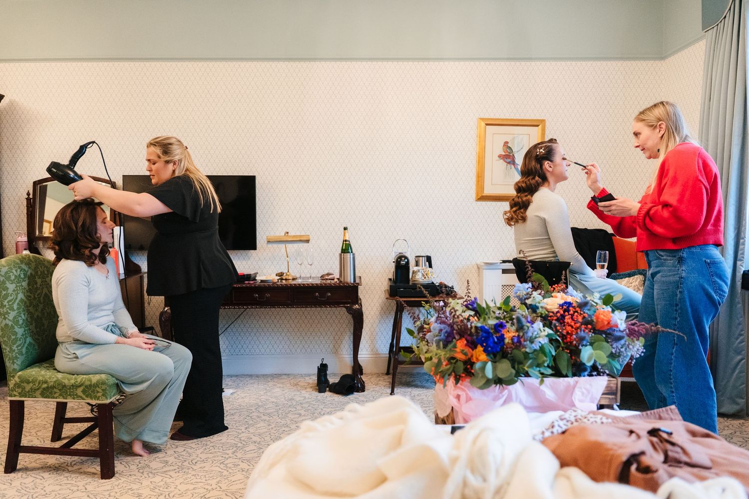 Wedding preparation scene with stylists doing hair and makeup while colorful flowers rest on nearby table.