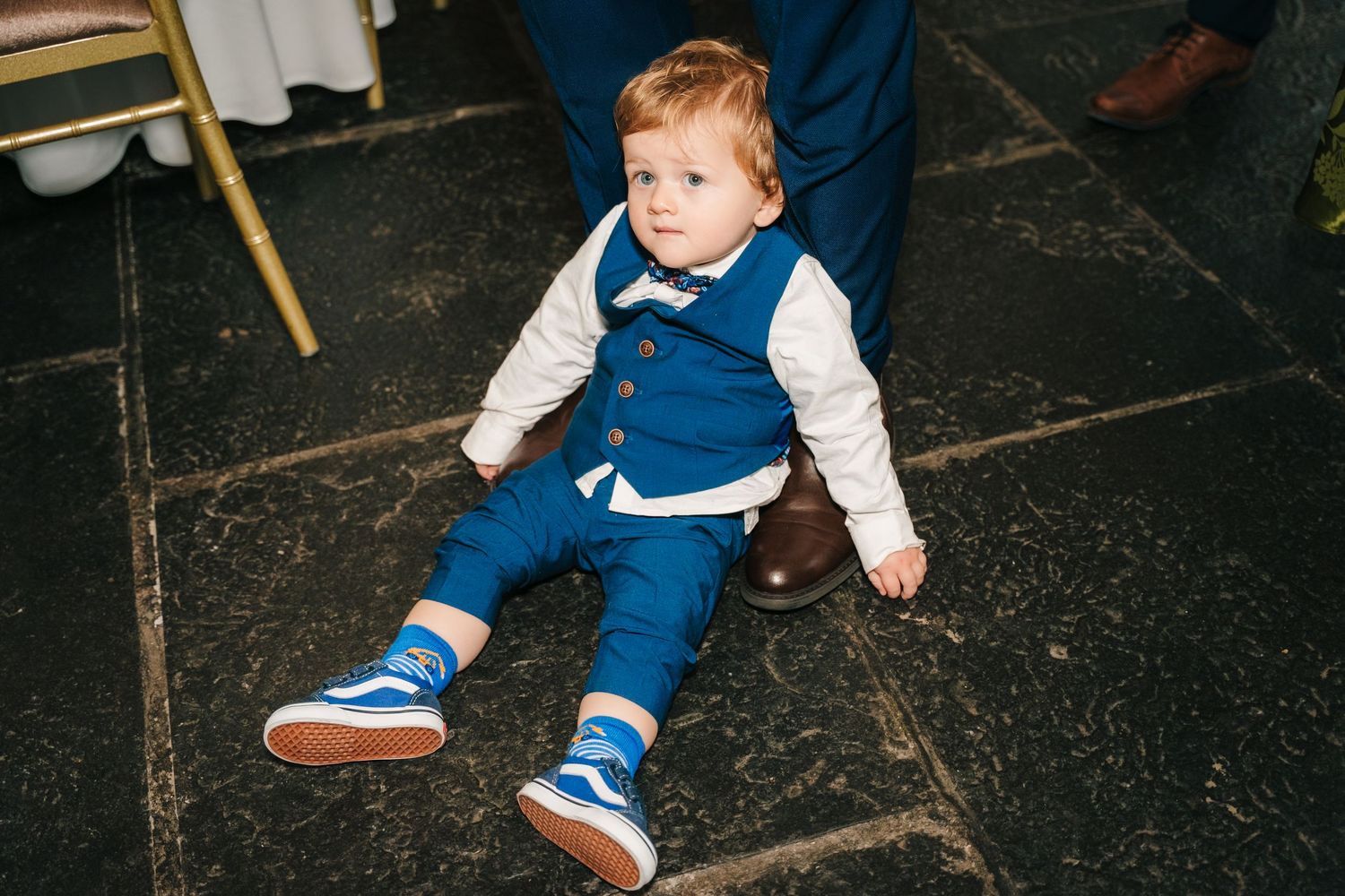 Young child in blue overalls sitting on dark stone floor at formal event.