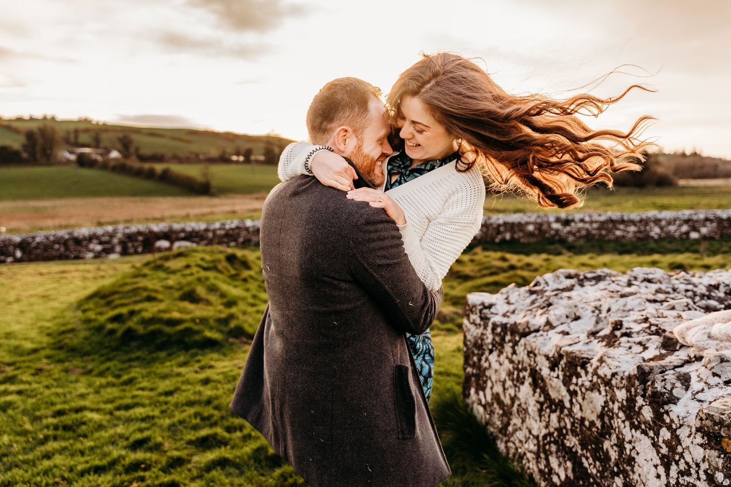 A couple shares a tender moment at sunset in an Irish countryside with stone walls and rolling green hills in the background. PHOTOGRAPHER D