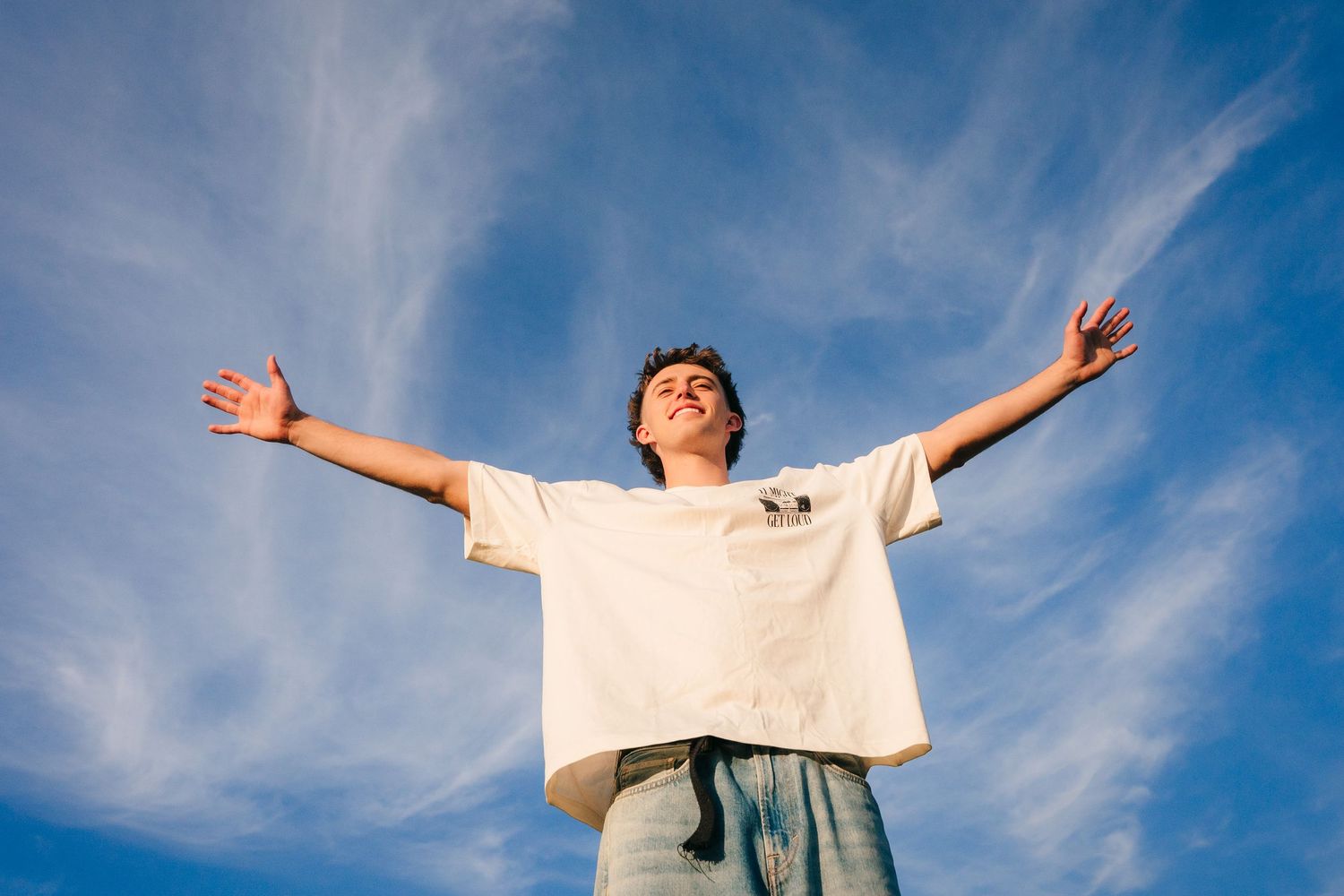 A person stands with arms outstretched against a bright blue sky with wispy white clouds. family PHOTOGRAPHER Dublin