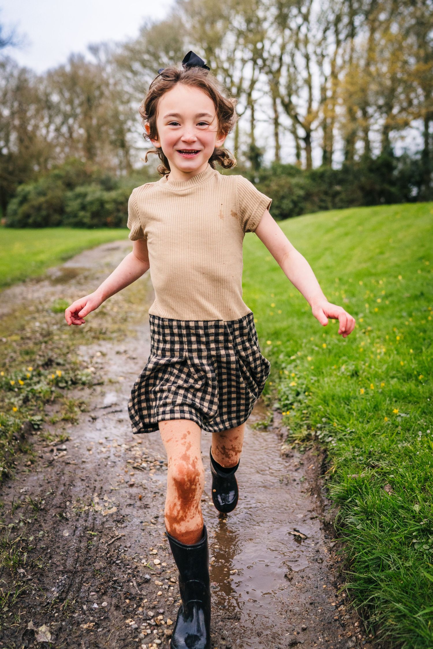 Energetic young person splashing through a muddy path wearing rain boots and a checkered skirt on a green hillside. family PHOTOGRAPHER Dubl