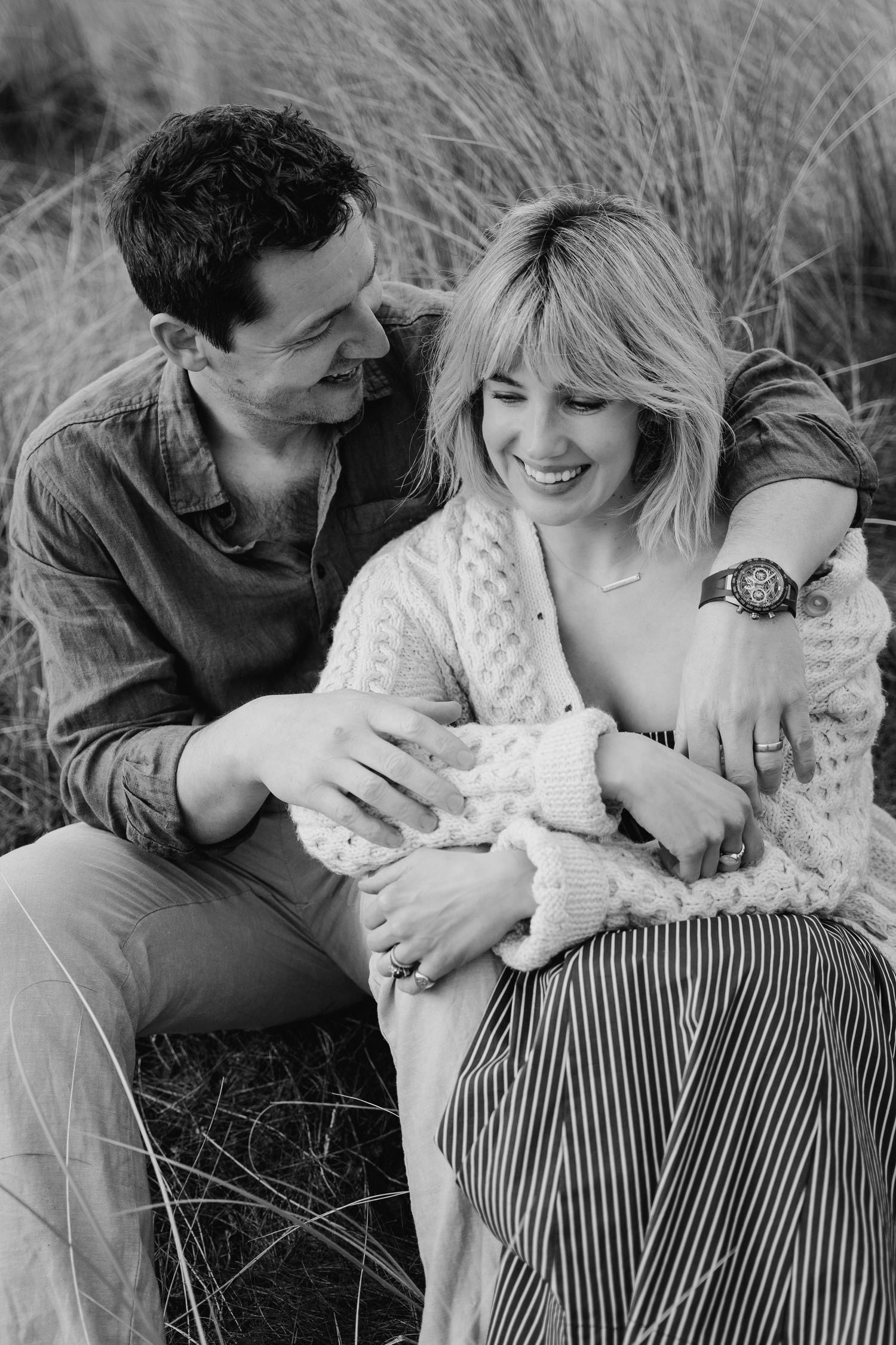 A couple sits in tall grass smiling while holding a newborn baby wrapped in white swaddling cloth. PHOTOGRAPHER Dublin ireland
