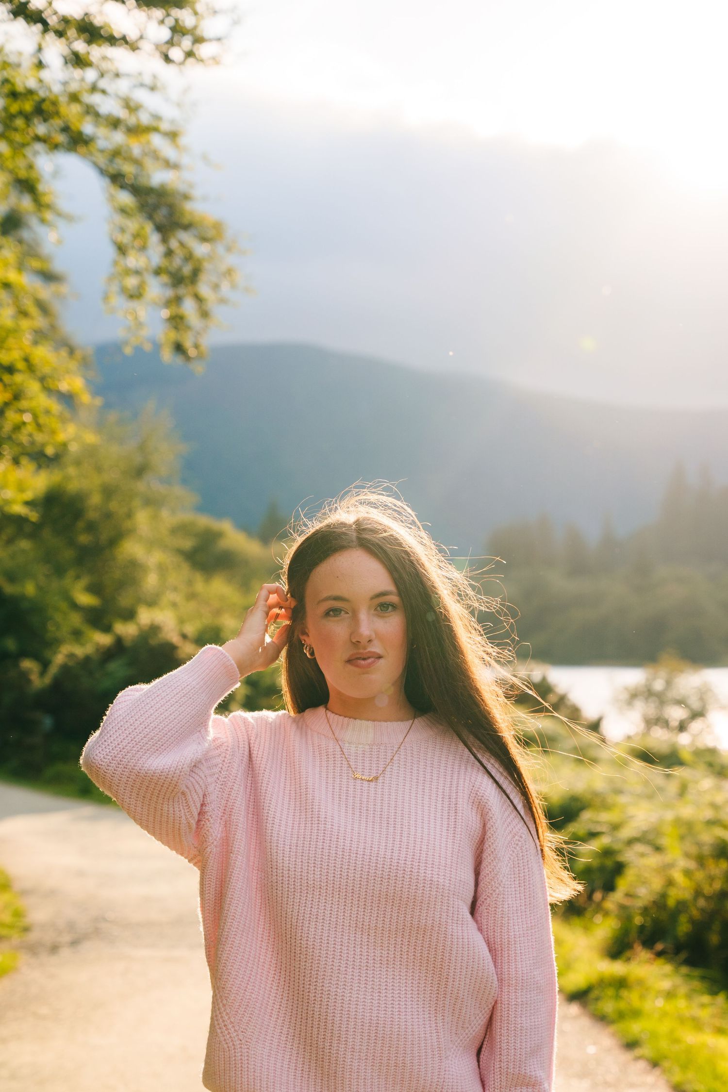 Family photographer Ireland Dublin A person in a pink sweater stands on a path with mountains and a lake in the background on a sunny day.