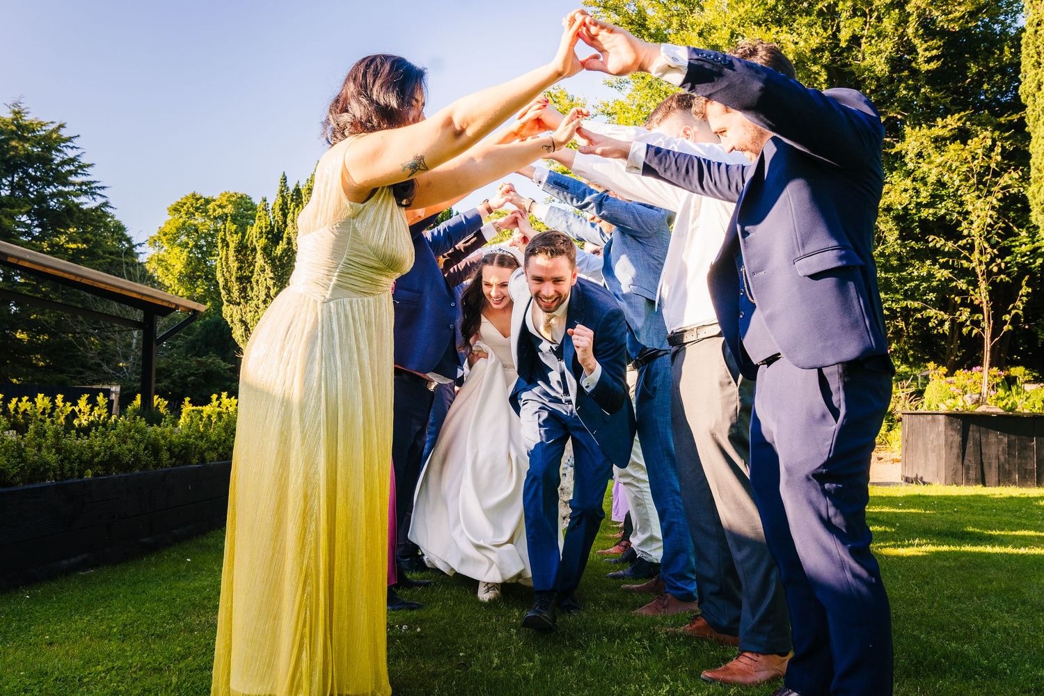 Poulaphouca Wedding photographer wicklow  Wedding party forms playful tunnel for celebration on sunny garden lawn.