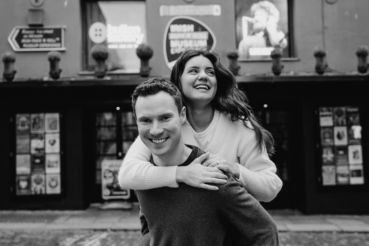Black and white photo of couple sharing playful piggyback ride outside storefront. Surprise proposal dublin photography