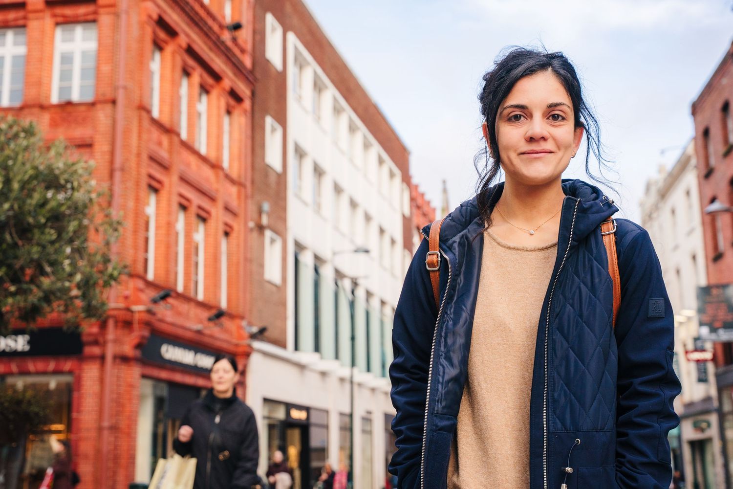 Someone wearing a navy quilted jacket and beige sweater smiles while standing on a street with red brick buildings. professional headshots